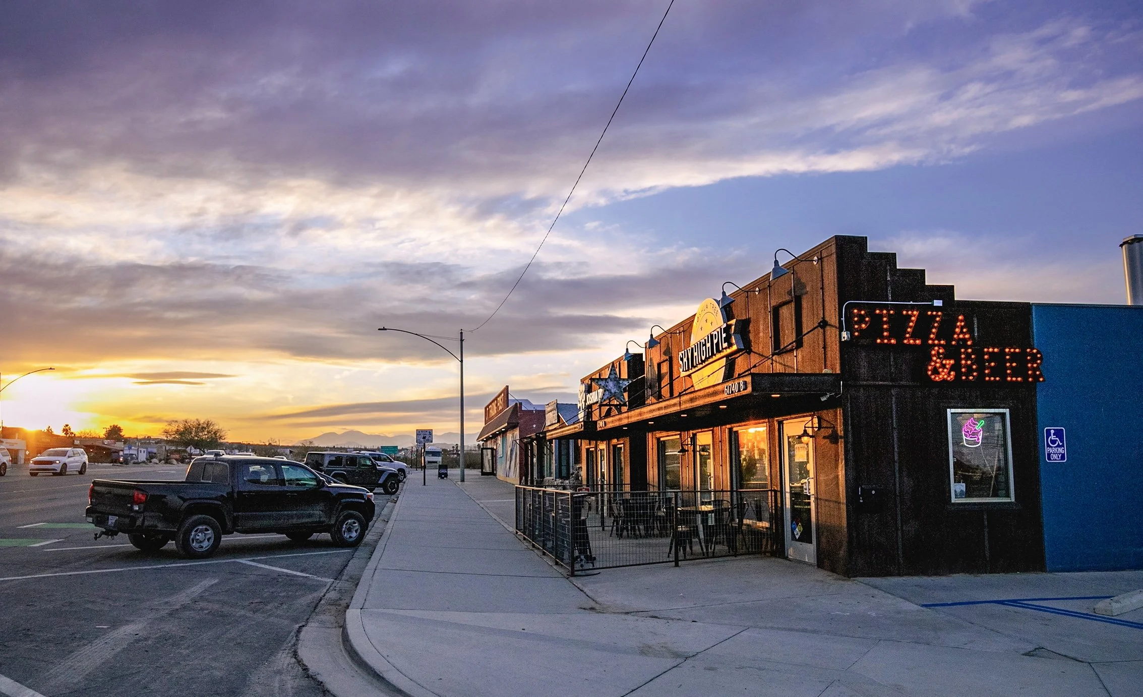 A strip mall at sunset with a restaurant that has outdoor seating, a neon sign indicating pizza and beer, parked cars, and a partly cloudy sky with the sun setting on the horizon.