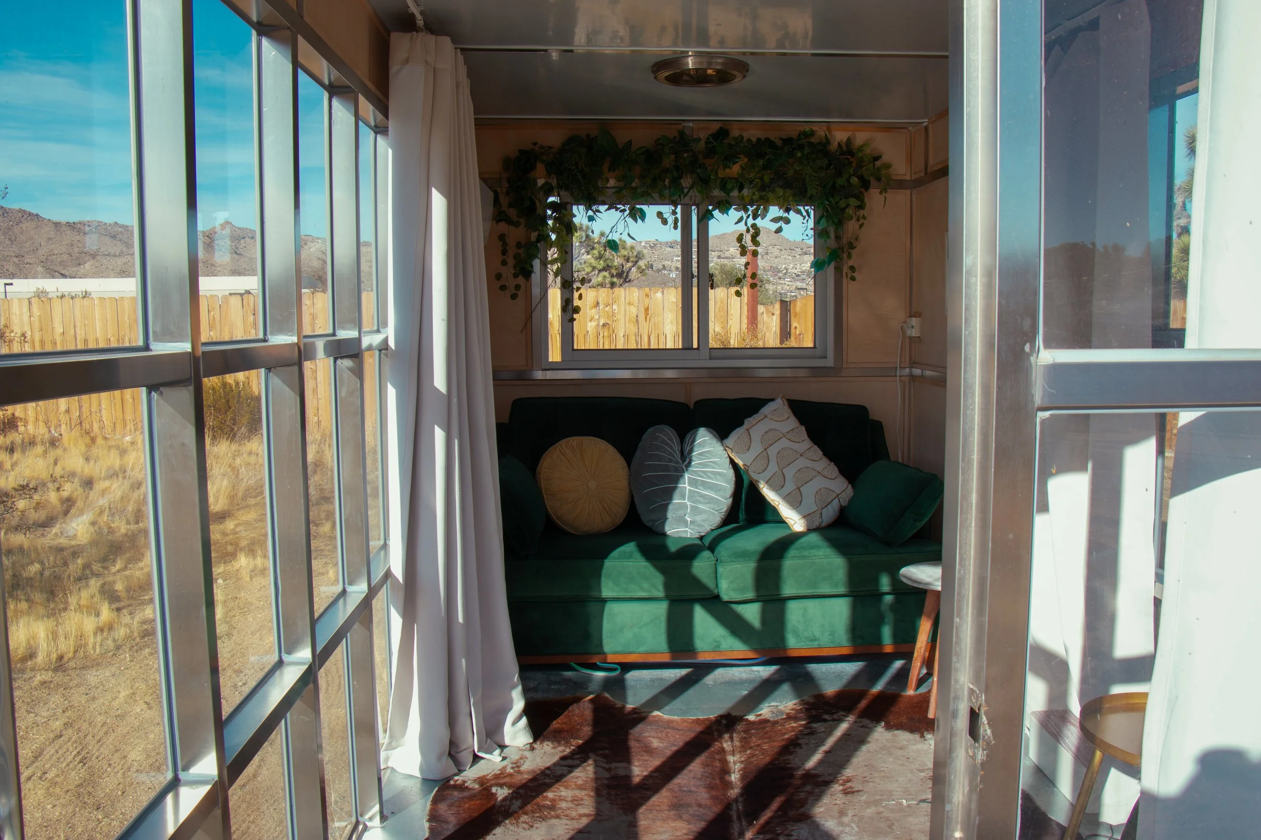 View from inside a small room or porch with large windows overlooking a desert landscape with mountains. The room has a dark green couch with assorted pillows, a cowhide rug, and hanging green plants. The space appears cozy and rustic with natural li
