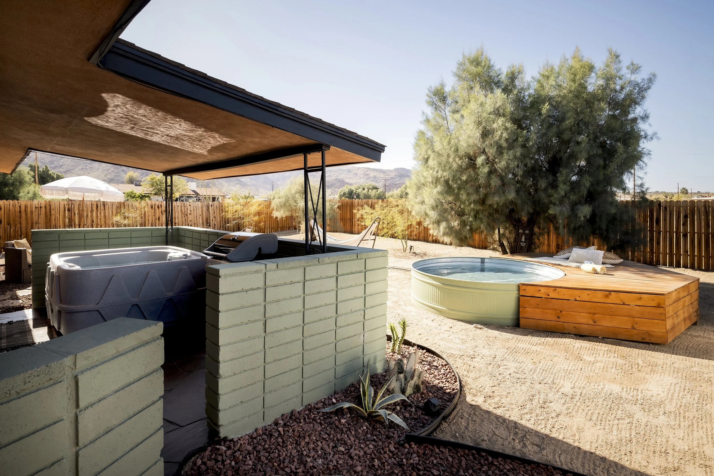 A backyard scene featuring a covered patio with a hot tub, a small round pool, and various outdoor furniture. There are trees, a wooden fence, and mountains in the background.