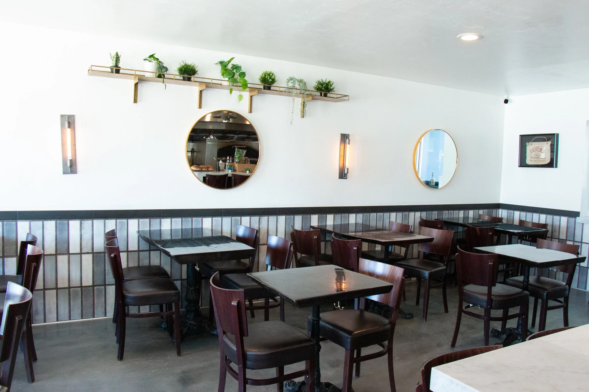 Inside of a restaurant with multiple wooden tables and chairs, decorative mirrors on white walls, and a high ceiling with simple lighting fixtures. There is a shelf with potted plants on the upper wall.