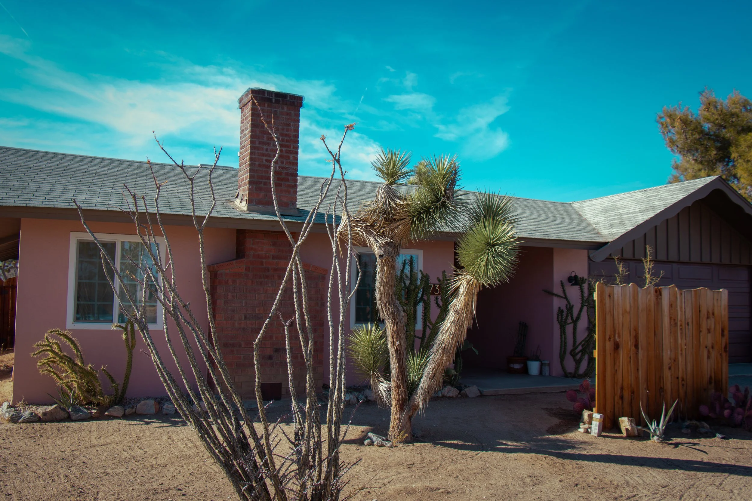 A house with a pink exterior, a brick chimney, and desert plants including a cactus and yucca plants in the front yard, under a blue sky with some clouds.