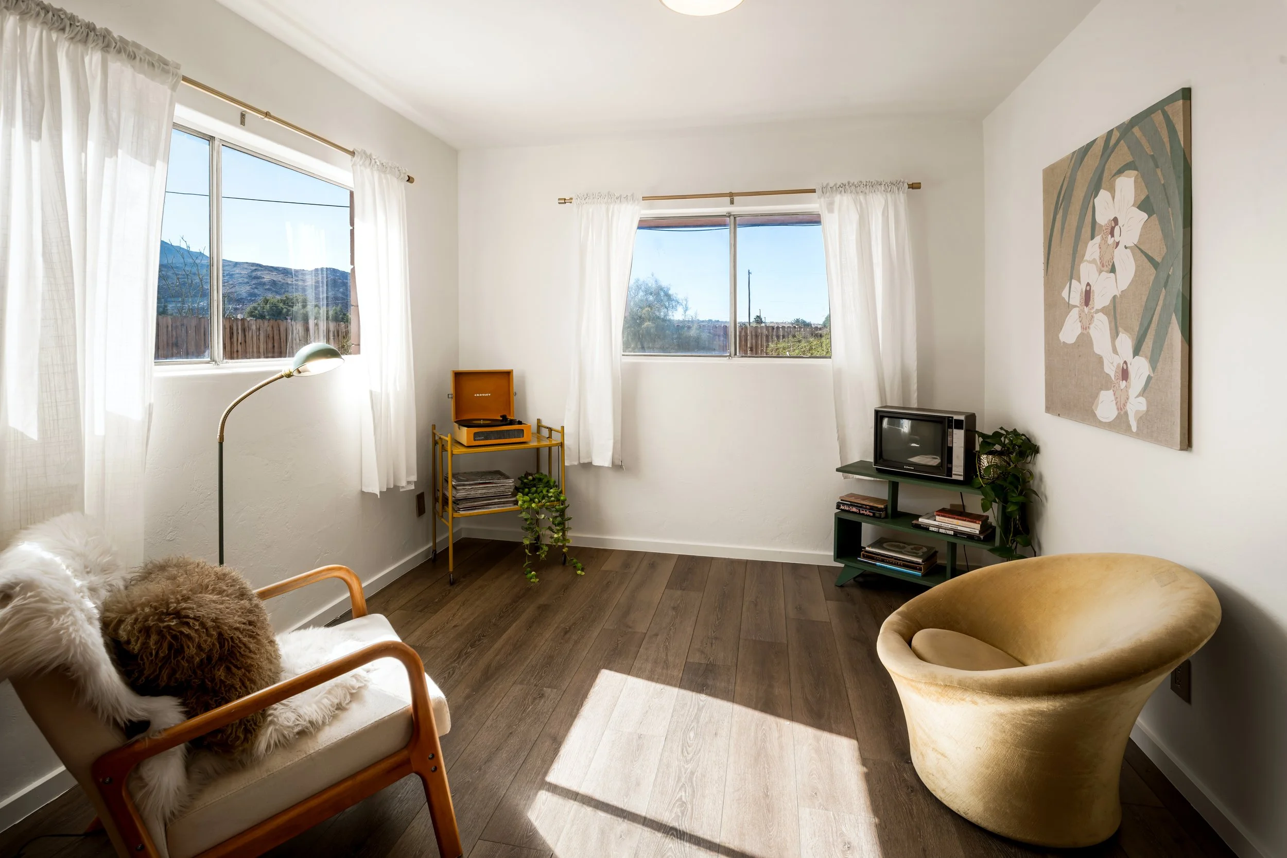 Living room with large windows, white curtains, a vintage record player on a yellow stand, a small TV on a green stand, wall art of white orchids, and plush stuffed animals on a chair.