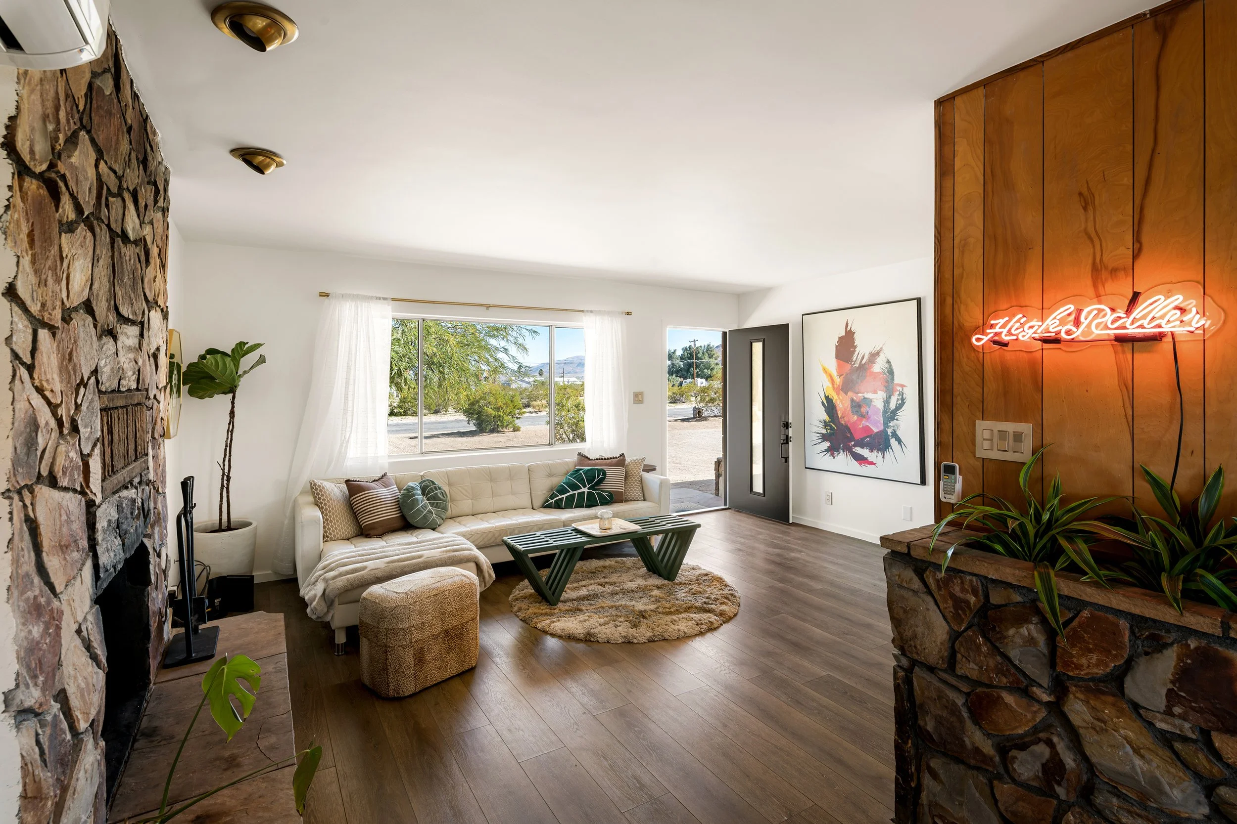 Living room with white walls, large window with white curtains, wooden floor, stone and wood accent walls, beige sofa with pillows, small coffee table, potted plants, colorful abstract painting, neon sign that says 'Hello Yall.'