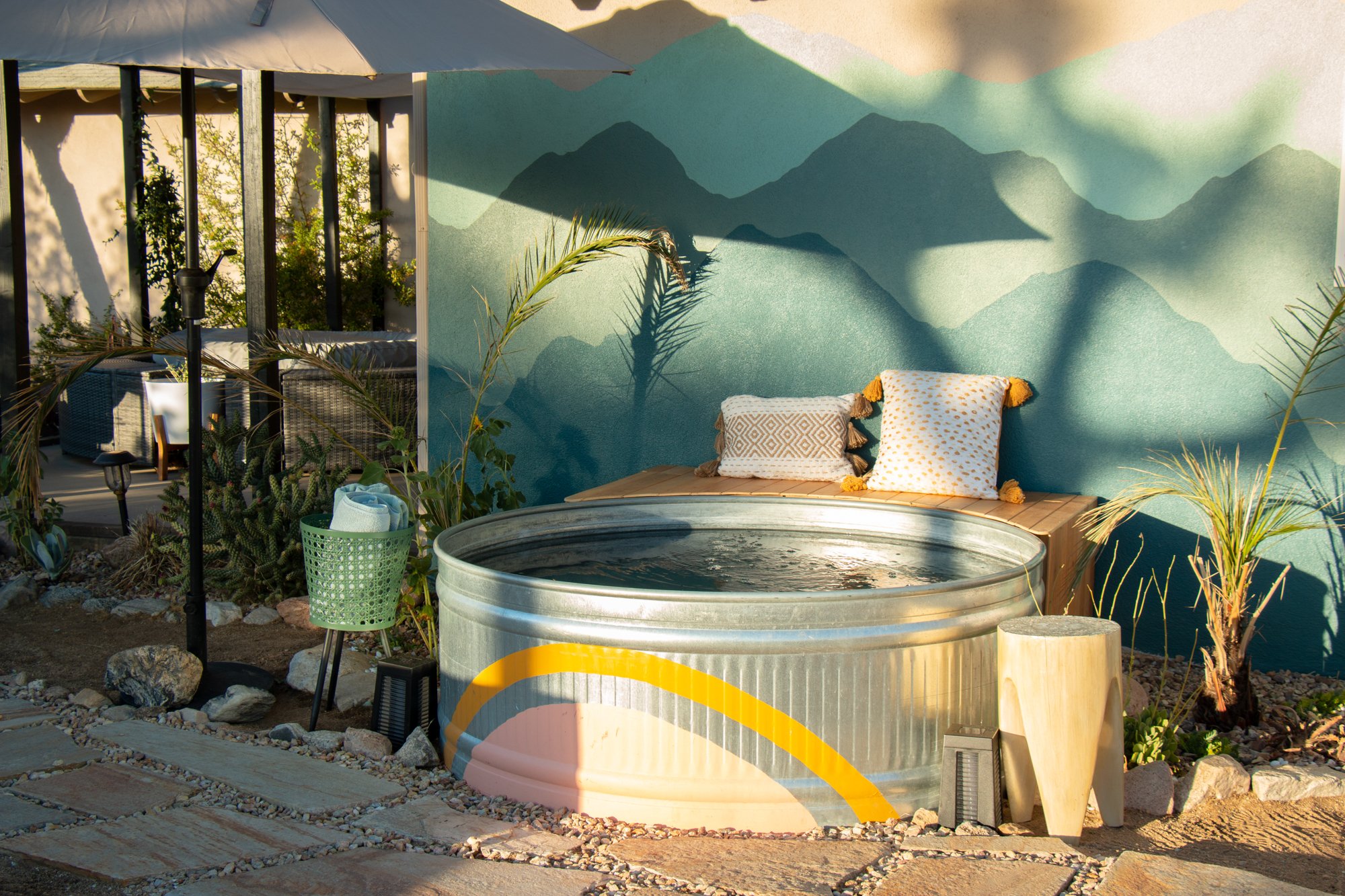 Outdoor patio with a painted mountain scene on the wall, featuring a hot tub, assorted pillows, greenery, and a partial umbrella.