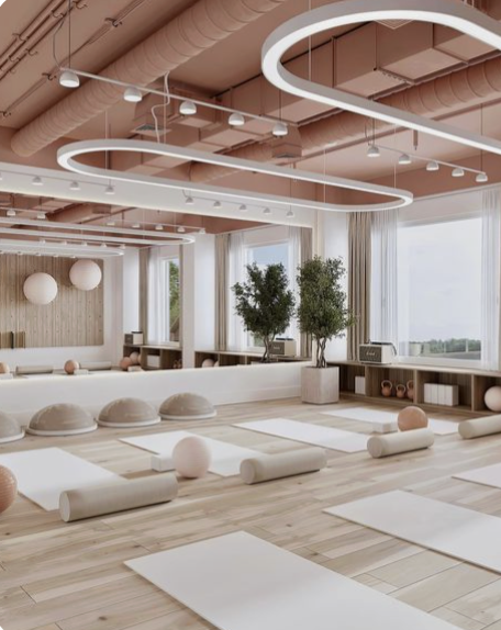 Bright yoga studio with wooden floors, large windows, and soft pastel decor, featuring yoga mats, bolsters, and two potted trees.
