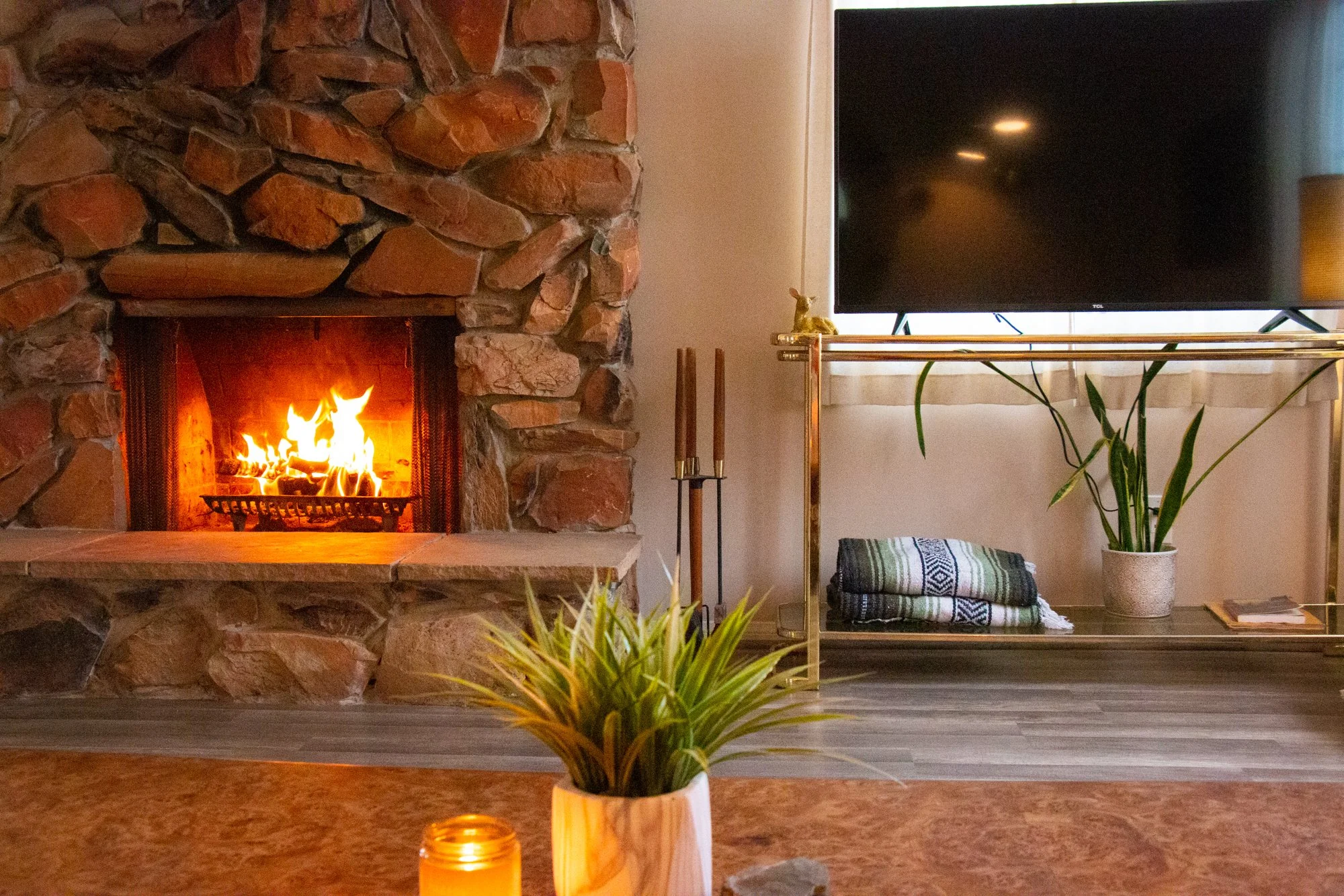 Living room with a brick fireplace, lit with a fire, and a flat-screen TV on a stand next to it. There are plants and decorative objects around, including a potted plant in the foreground, the floor is wood, and there are neatly folded throws or blan
