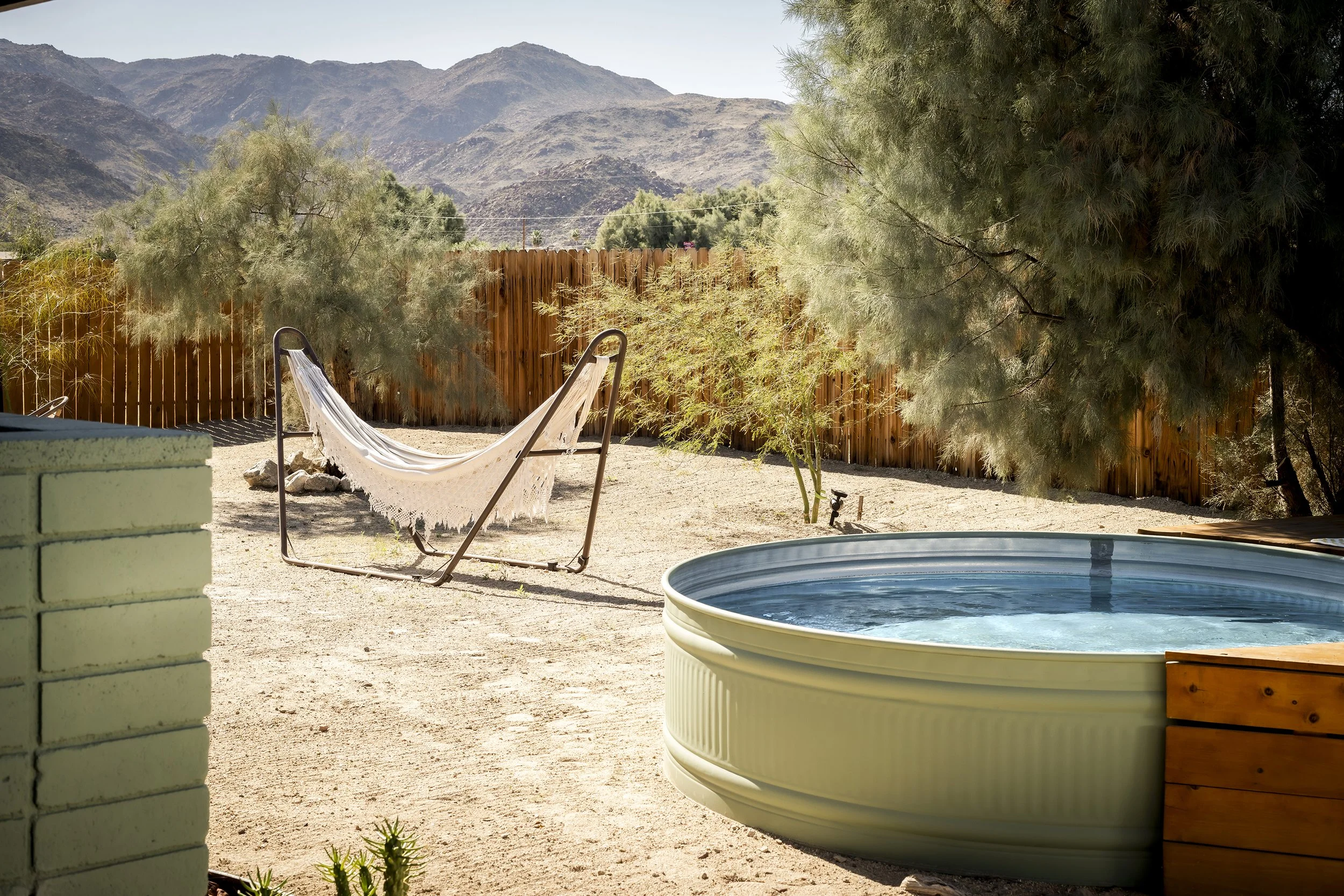 A backyard scene with a hot tub, a hammock, and trees, with mountains in the background on a sunny day.