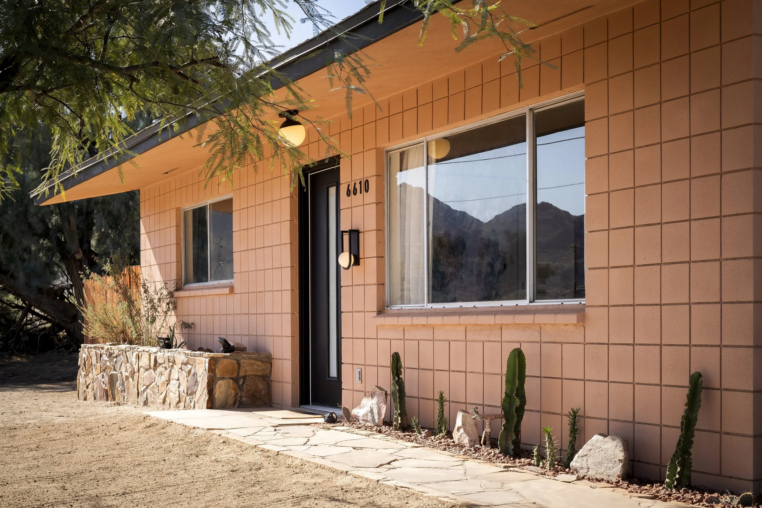 A modern house with peach-colored brick walls, large windows, and a black front door. The house has a stone pathway front and desert plants including cacti. A tree extends over the house, with mountains visible in the background.