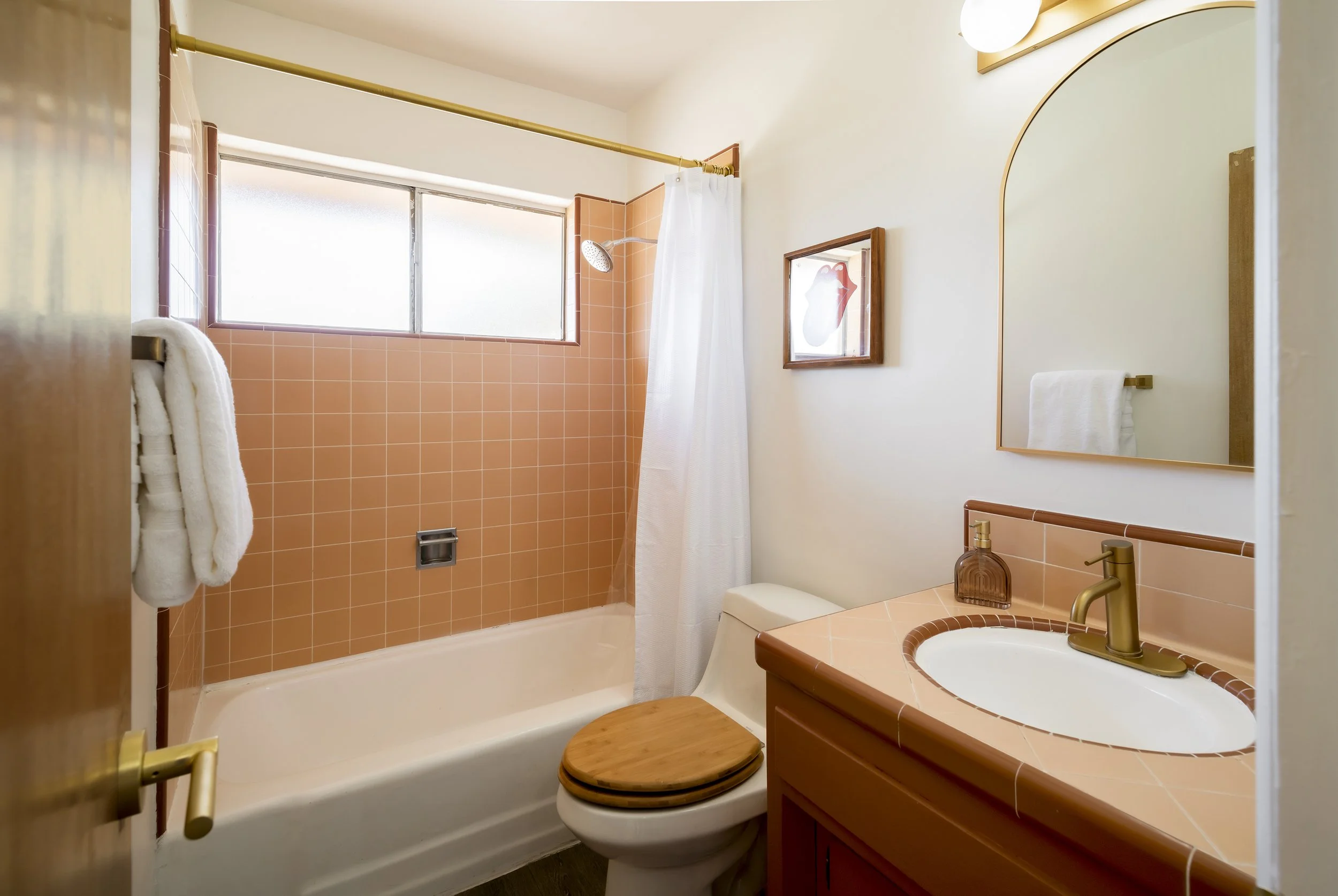 Bathroom with pink tiles on the shower wall, a showerhead, frosted window, mirror, towel rack with white towels, wooden toilet seat, and a sink with a matching pink tile countertop and gold fixtures.