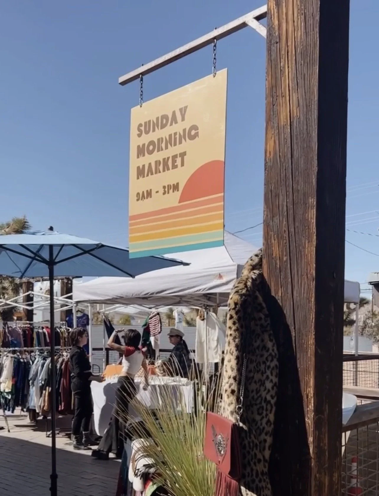 People shopping at an outdoor Sunday morning market with clothing stalls and vendors under tents on a sunny day.