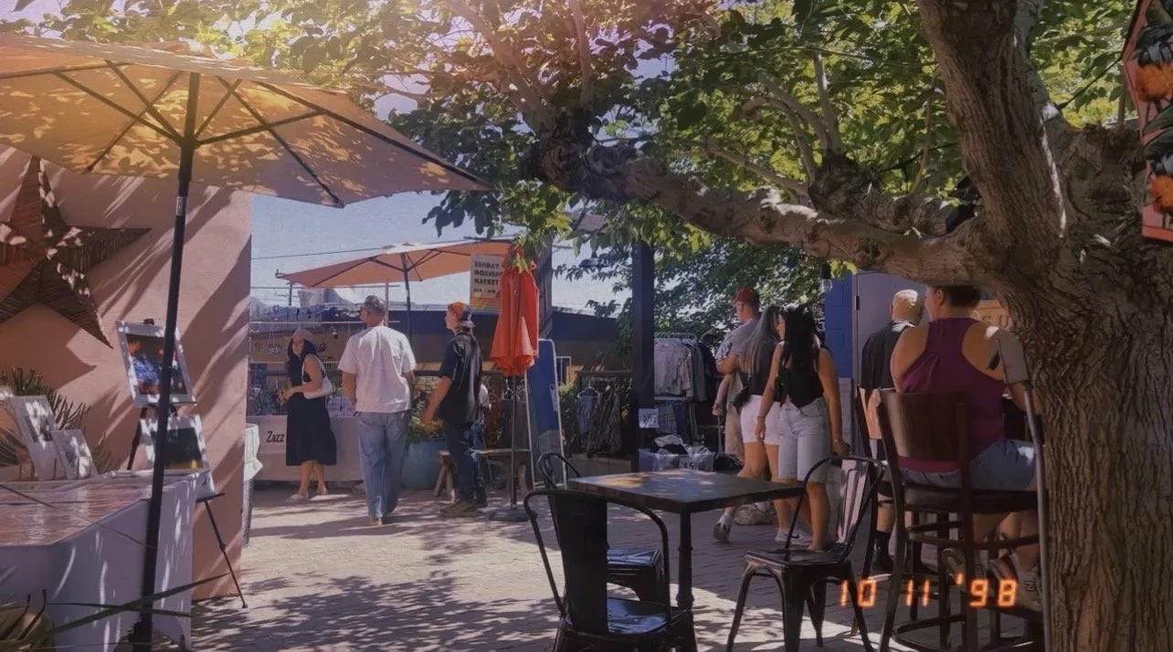 People browsing an outdoor market under large umbrellas with a tree in the foreground and a clear blue sky in the background