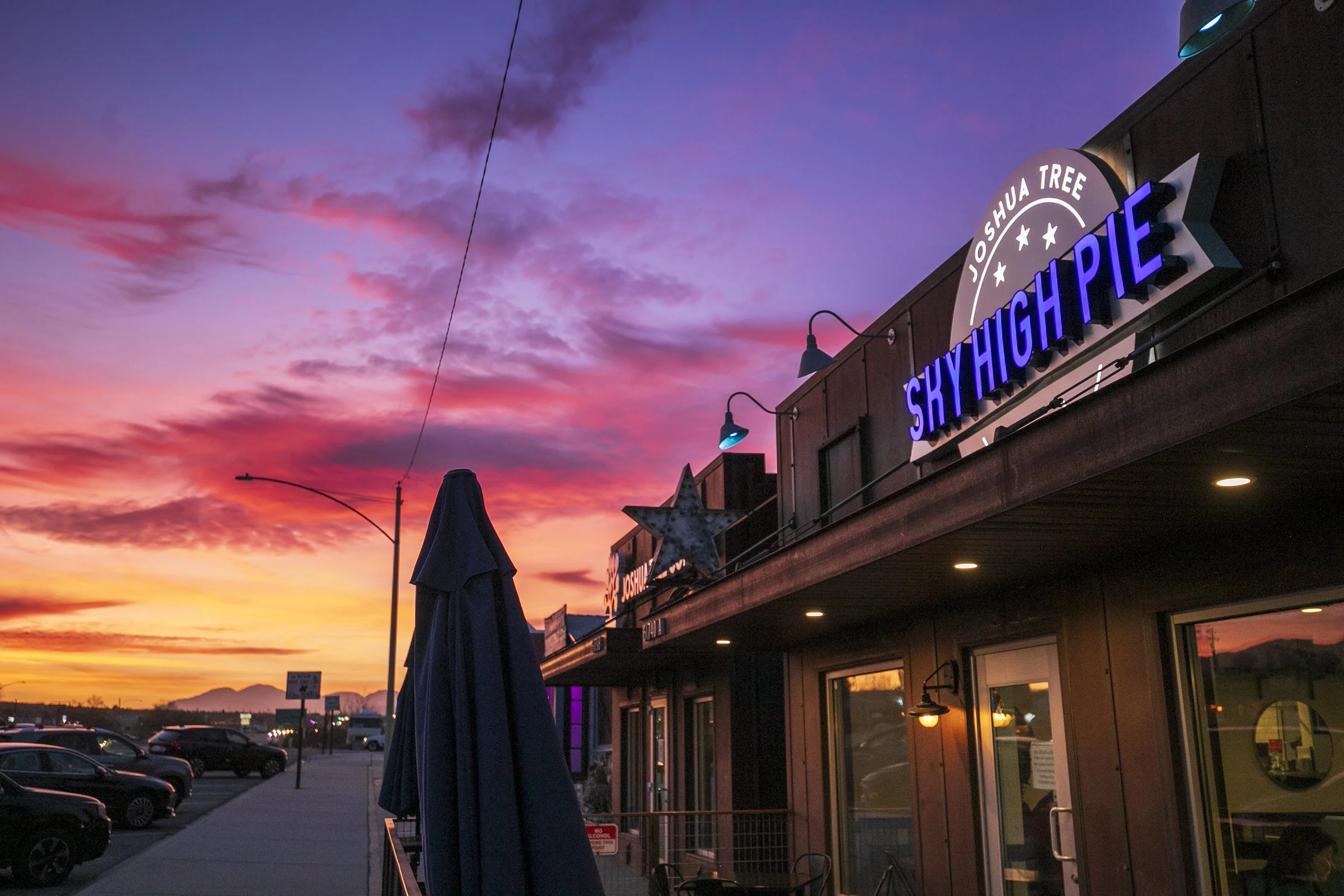 View of a storefront with illuminated sign reading "Joshua Tree Sky High Apple" against a colorful sunset sky, with parked cars and a few street lights visible in the background.