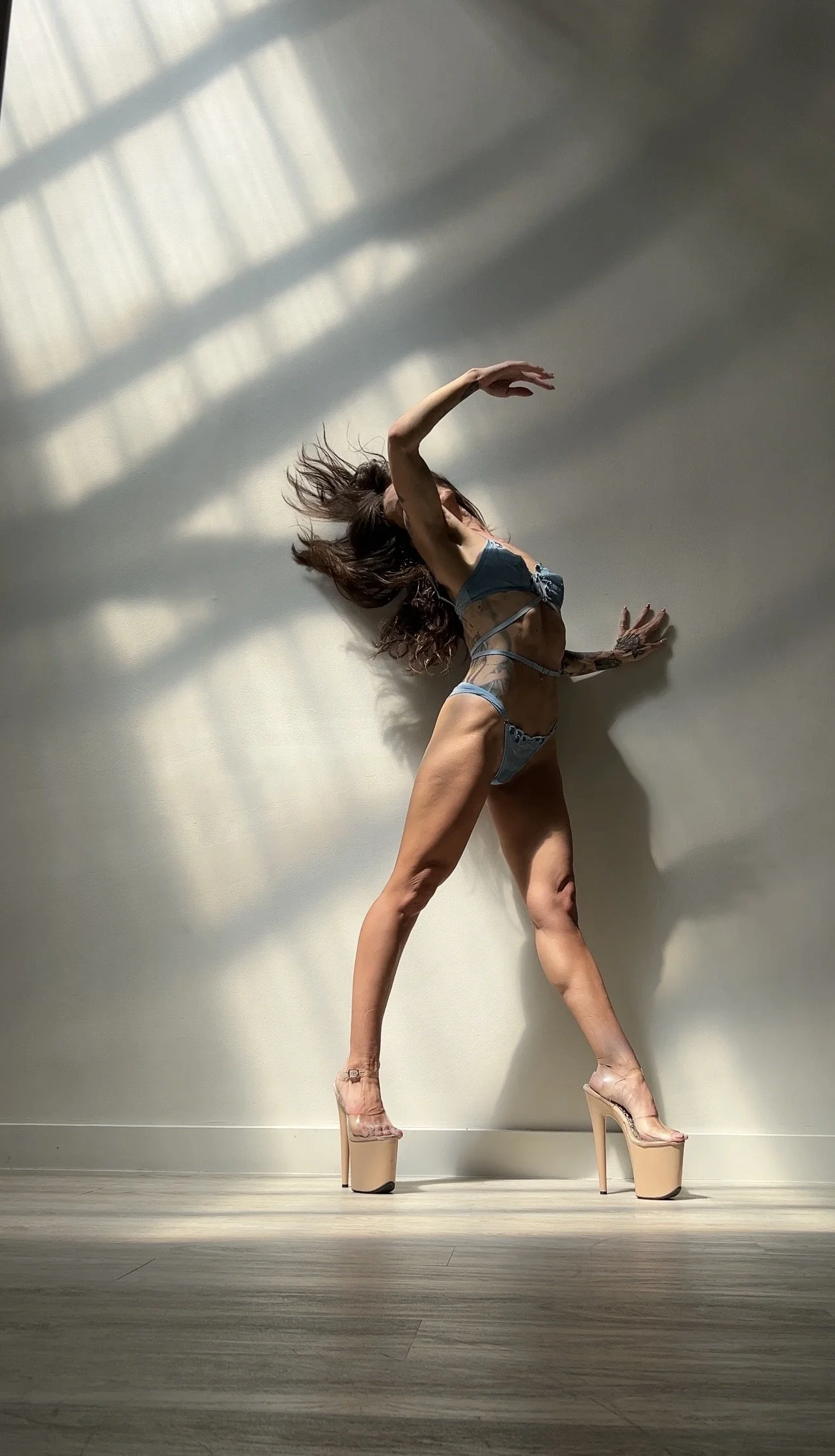 A woman in lingerie wearing high heels, posing against a wall with shadows from window blinds, with her head tilted back and her hair flowing.