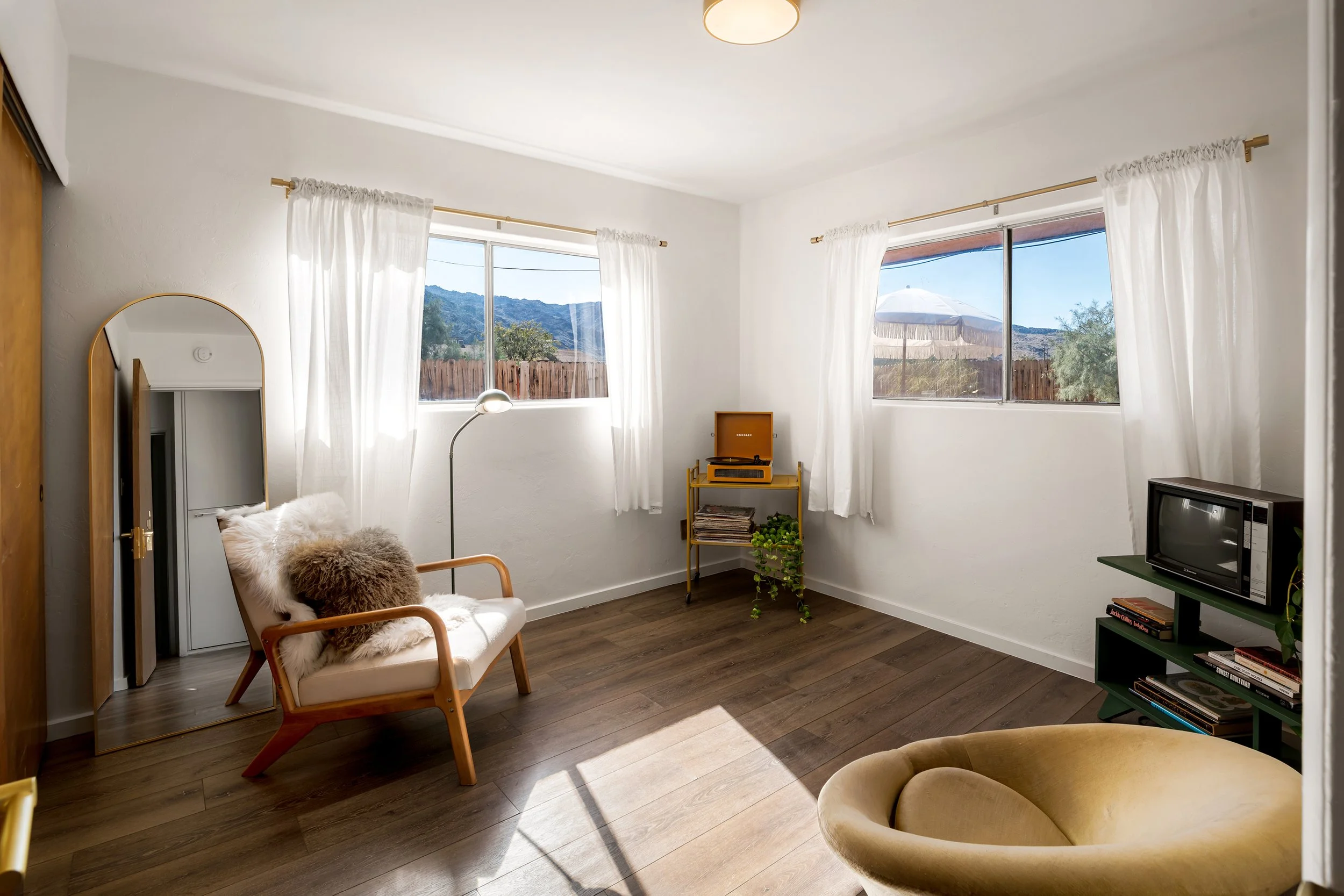 A bright living room with white walls and dark wood flooring, featuring a cozy chair with a fur throw, a small teal bookshelf with magazines and plants, a vintage yellow record player, a standing lamp, and large windows with white curtains overlookin