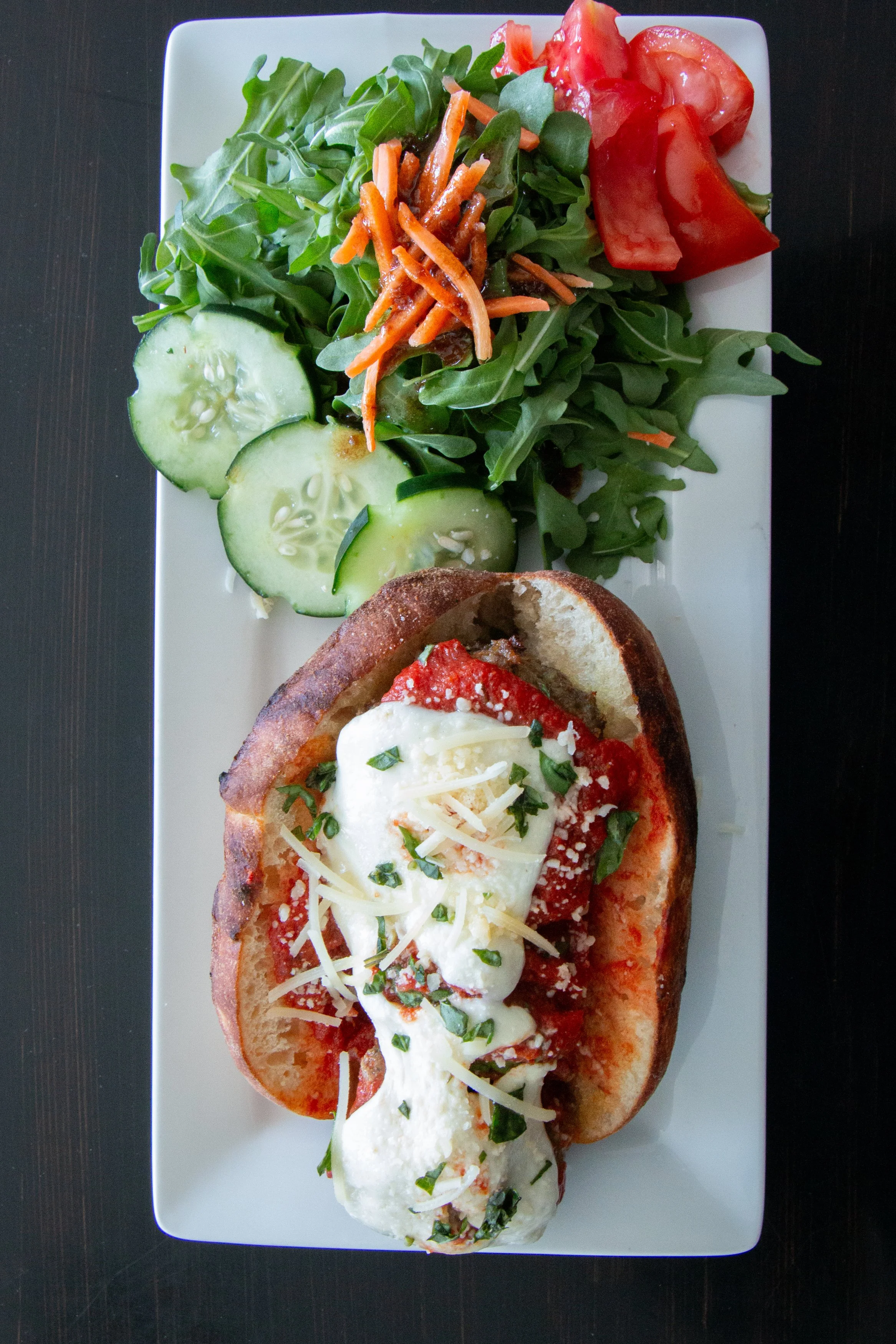 A rectangular white plate with a garden salad consisting of arugula, cucumber slices, shredded carrots, and tomato wedges, alongside a small pizza topped with mozzarella cheese, chopped herbs, tomato sauce, and grated cheese.