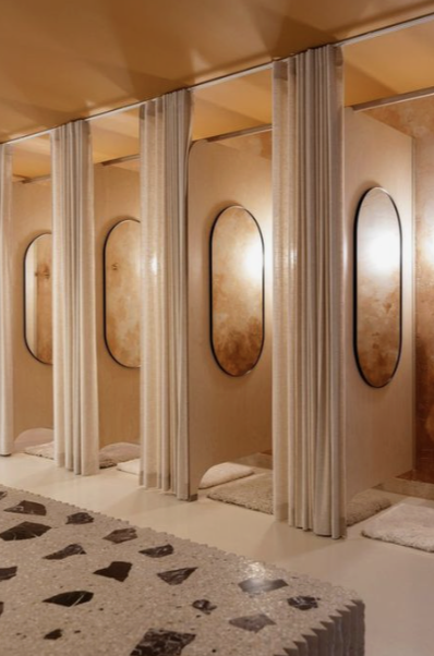 A row of private salon or spa booths with beige curtains, padded seats, and oval mirrors on the beige walls.