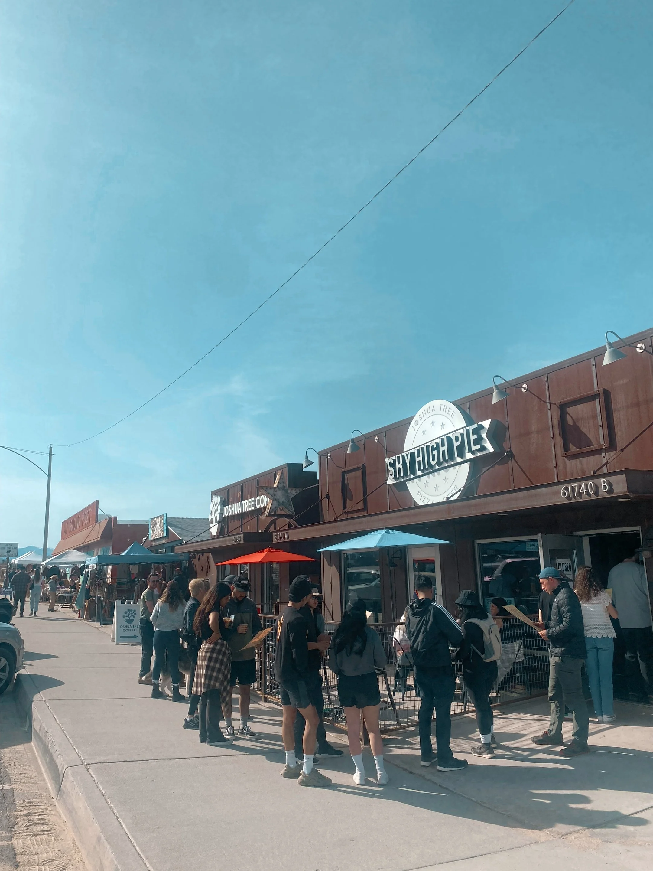 Crowd of people standing in line outside a restaurant called Sky High Pie, on a city sidewalk on a sunny day.