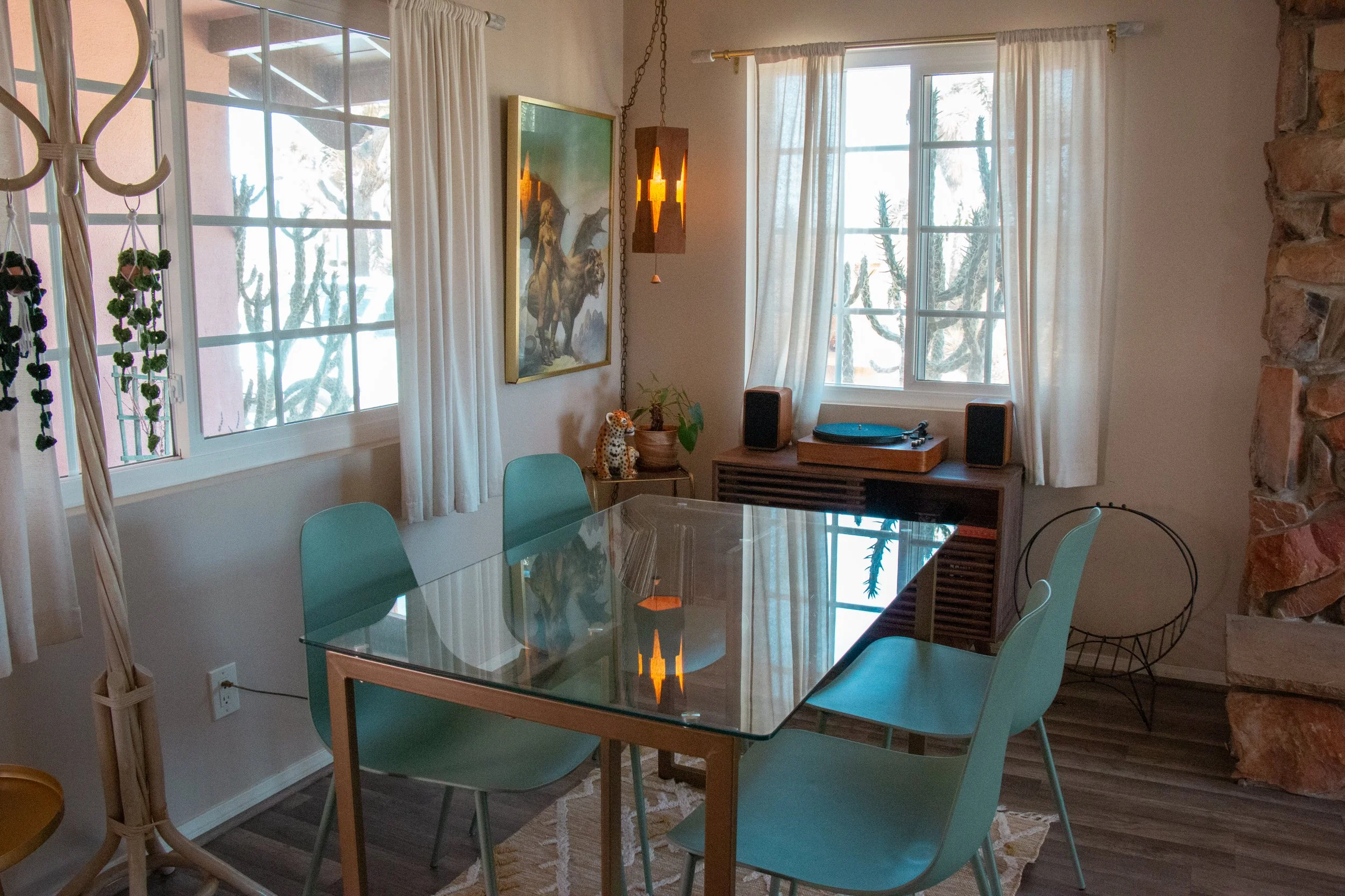 Bright dining room with large windows covered by white curtains, artwork featuring horses on the wall, a vintage turntable, and a ceramic leopard figurine on a side table. Tan leather chairs surround a glass dining table. A stone wall is visible on t