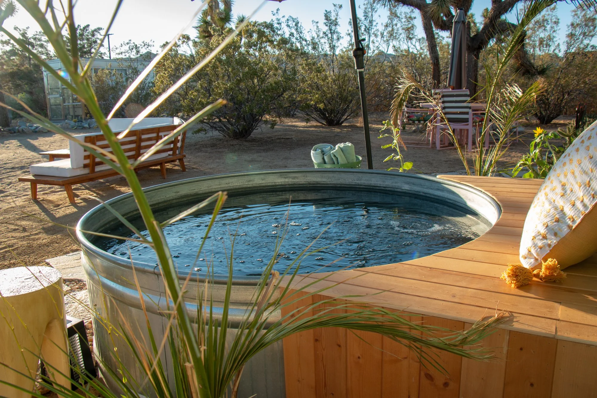 A backyard outdoor scene with a hot tub, lounge chairs, and a dining area surrounded by desert plants and trees, during sunset.