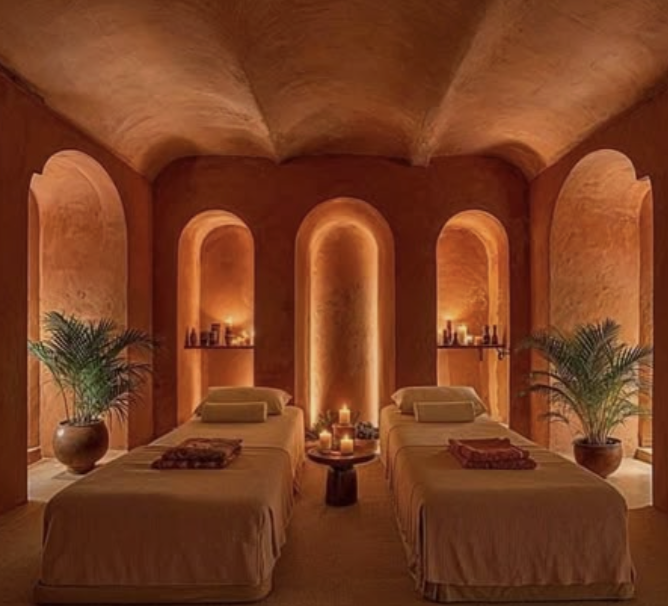 A relaxing spa room with two massage beds, candlelight, and potted plants, featuring warm, earth-tone walls and arched niches with small decorative bottles.