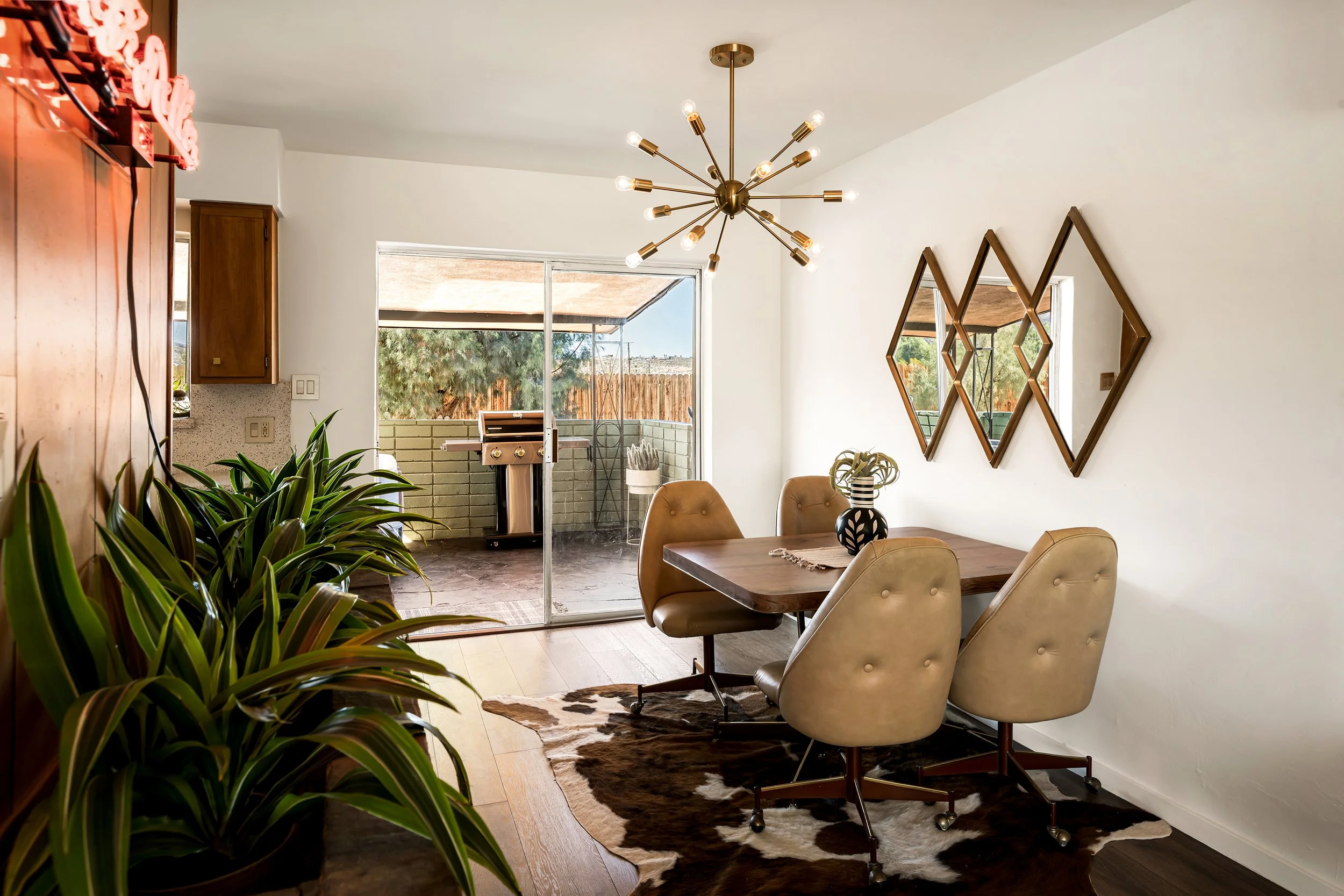 A dining room with a wooden table surrounded by light tan upholstered chairs. There is a black and white patterned vase on the table. A modern gold chandelier hangs from the ceiling, and three diamond-shaped wooden mirrors are on the white wall. To t