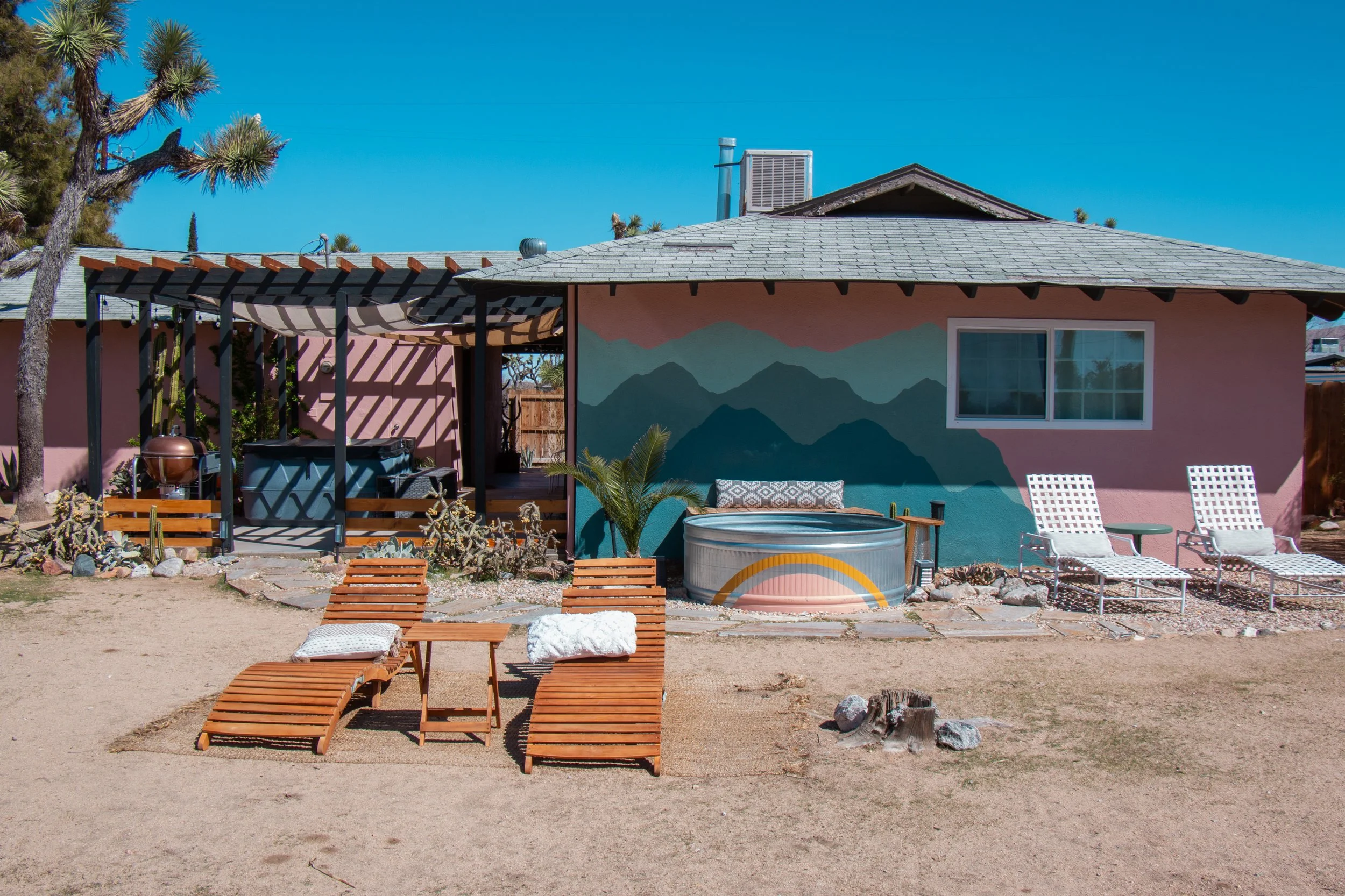 A backyard with wooden lounge chairs on sandy ground, a pink house with mountain mural, trees, and patio furniture under a pergola.