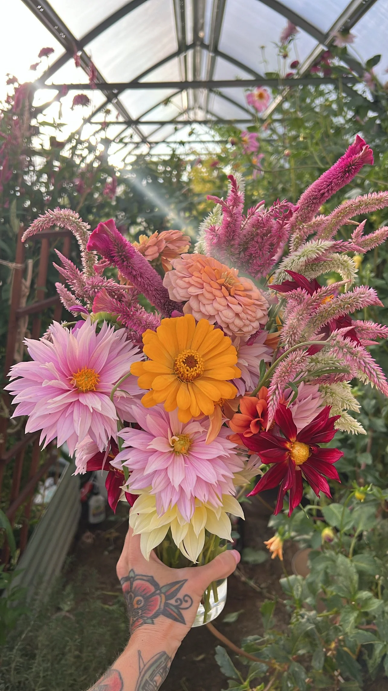 A person with tattoos on their hand holding a colorful bouquet of flowers in a greenhouse.