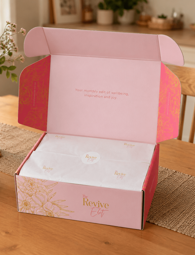 Open pink and gold box labeled "The Revive Edit" on a wooden table with white tissue paper inside and a message that reads, "Your monthly edit of wellbeing, inspiration and joy."