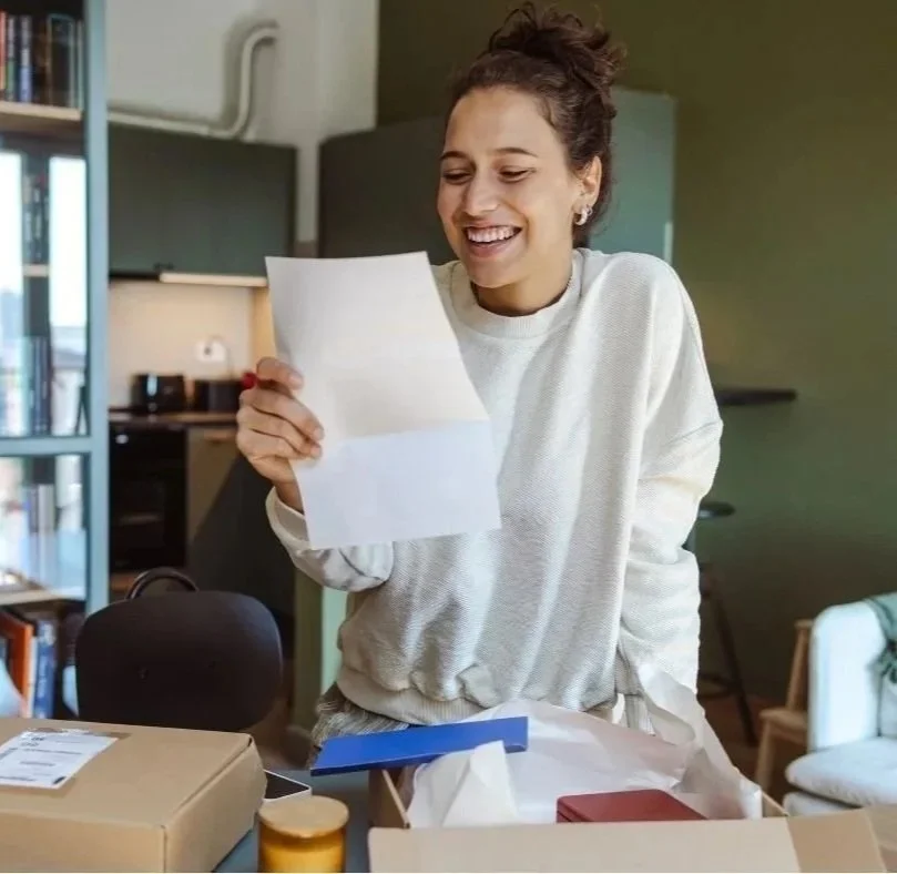 A woman smiling while reading a letter in a cozy, modern kitchen or living space with bookshelves and warm lighting.