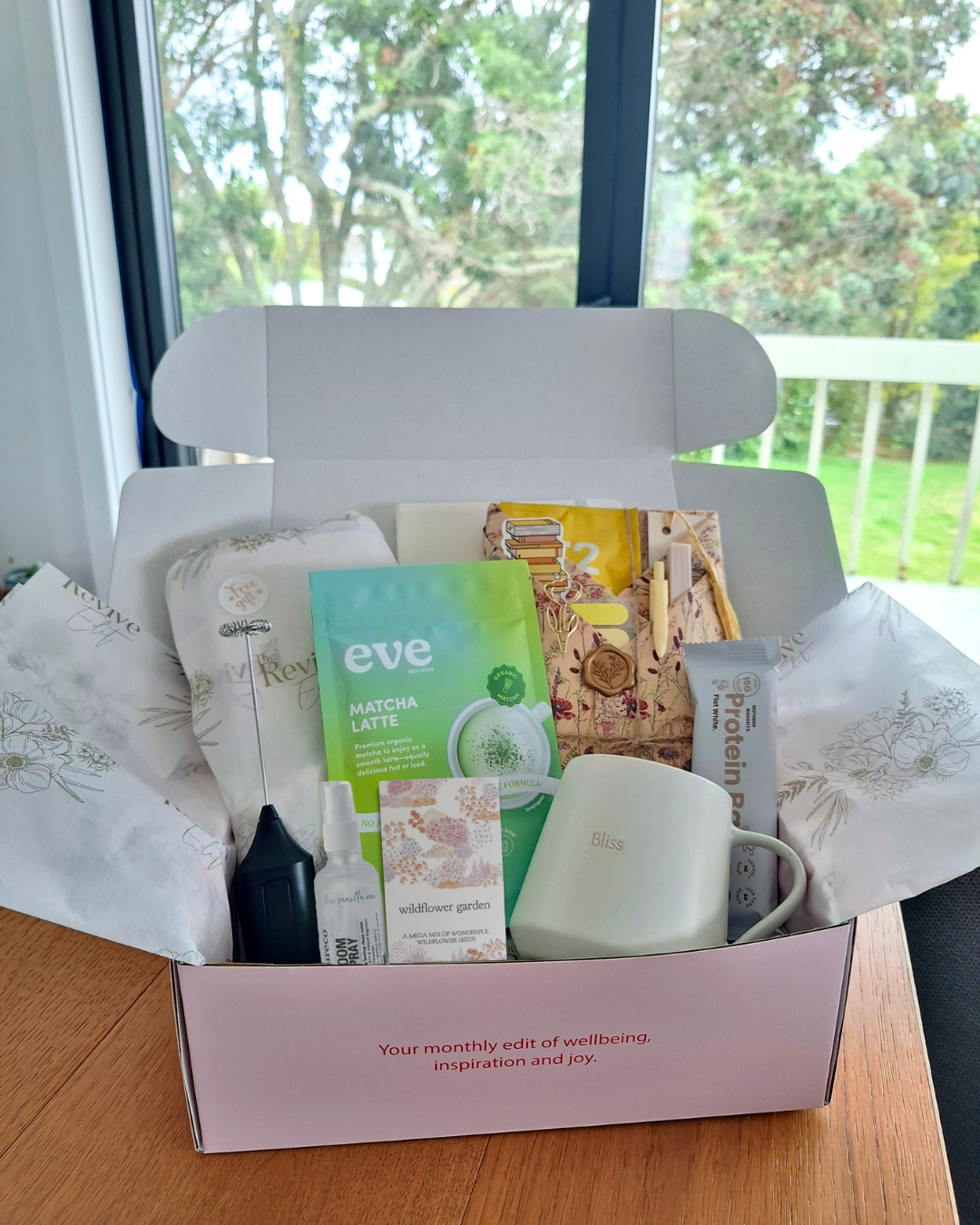 A Revive Edit box filled with various wellness and lifestyle items on a wooden table near a window with trees outside. Items include a white mug with the word 'Bliss,' a packet of Matcha Latte, a card labeled 'wildflower garden,' a small spray bottle