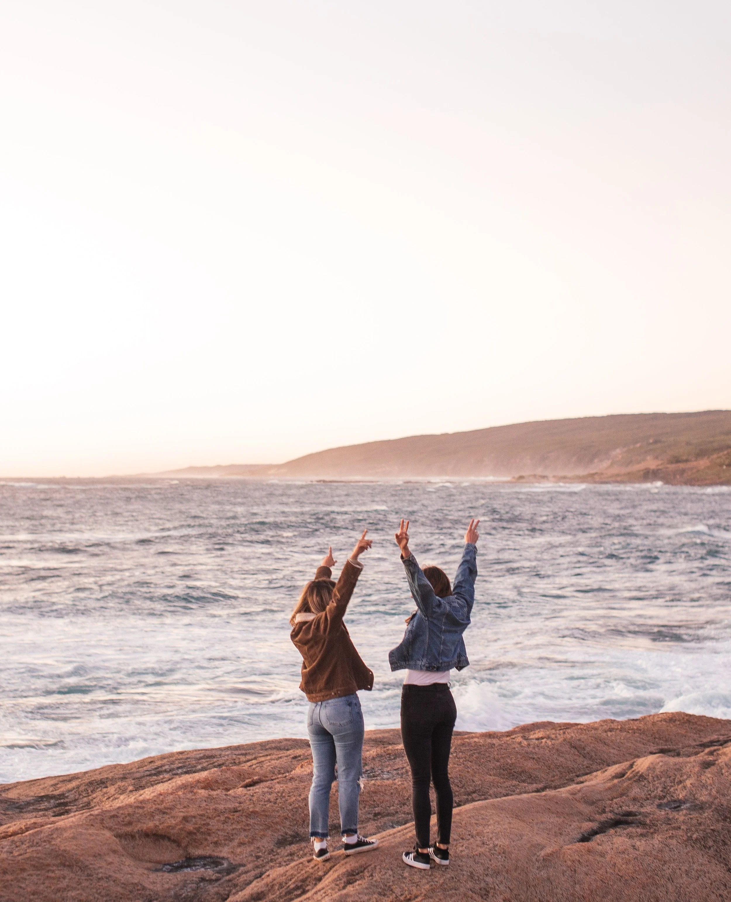 Two women standing on rocks at the beach during sunset, holding their arms up making peace signs, with the ocean and hills in the background.