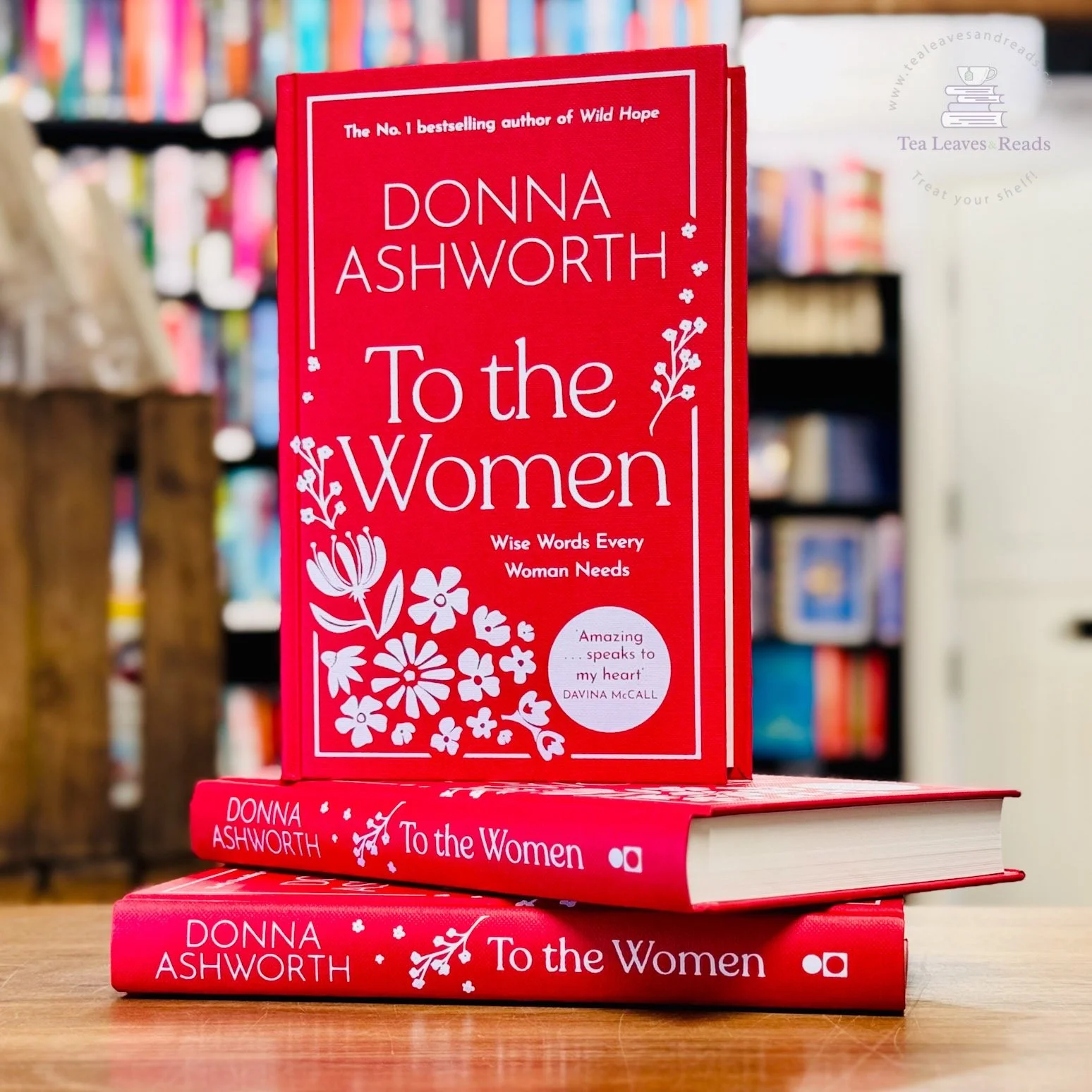 Stack of three red books titled "To the Women" by Donna Ashworth displayed on a wooden table in a bookstore, with bookshelves in the background.