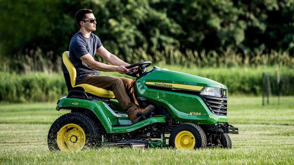A man wearing sunglasses drives a green and yellow John Deere X584 lawn tractor across a grassy field.