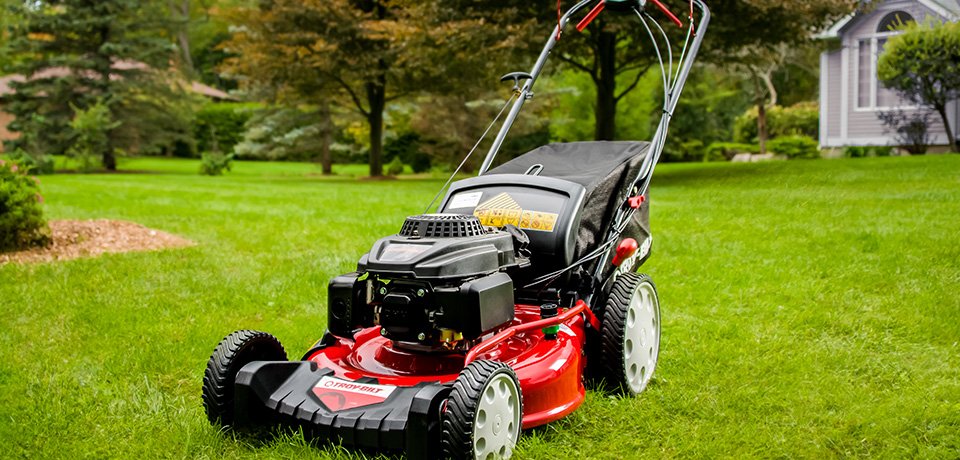 A red and black gas-powered lawn mower on a neatly maintained green lawn, with a house and trees in the background.