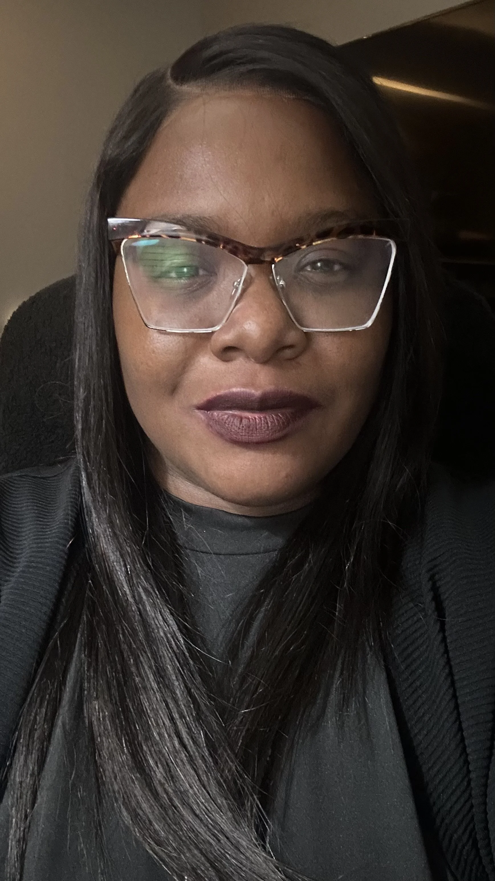 A woman with long black and gray hair, wearing large tortoiseshell glasses, smiling with dark lipstick, dressed in black, sitting indoors with a dark background.