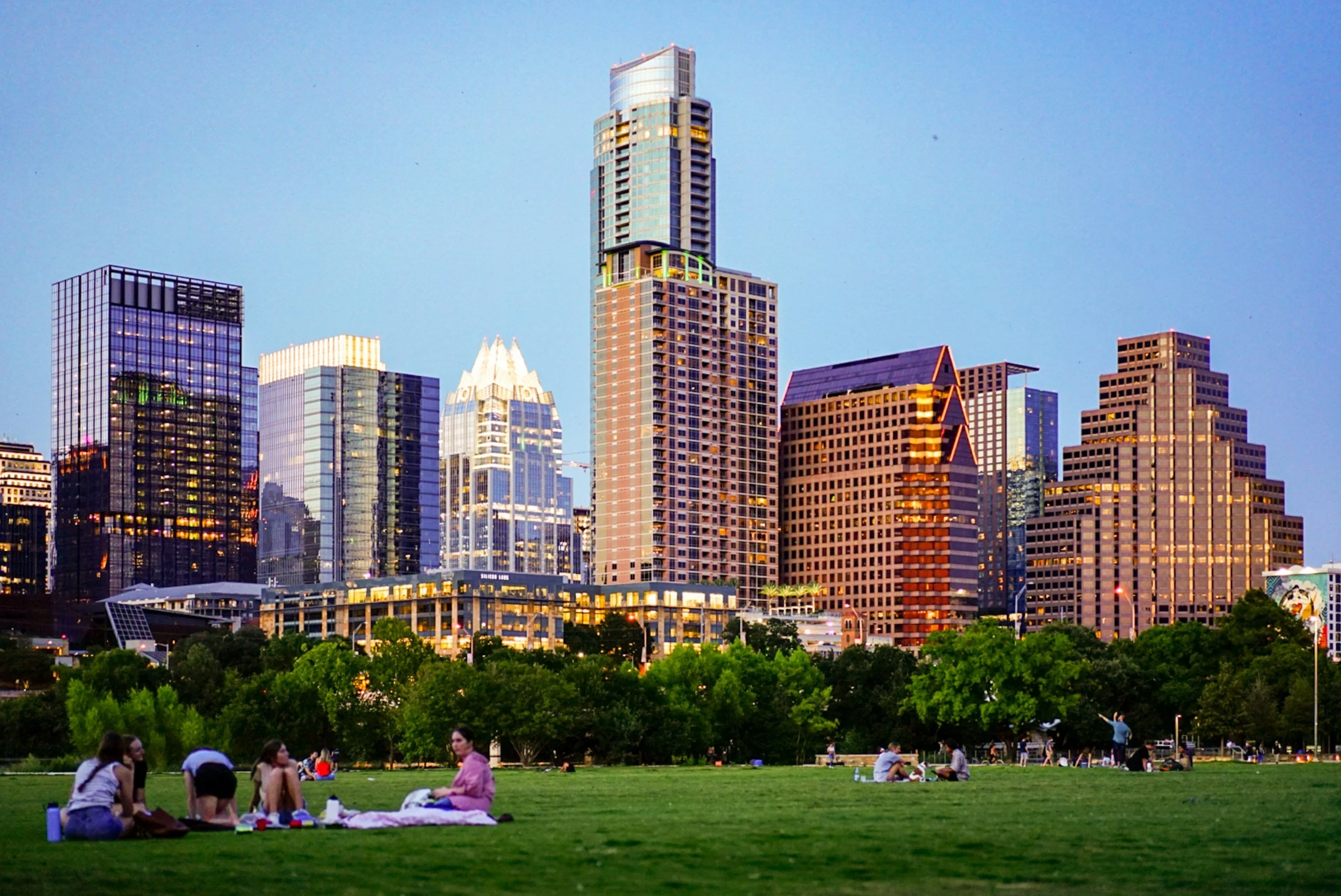 People sitting on the grass in a park with a city skyline featuring tall buildings in the background during evening.