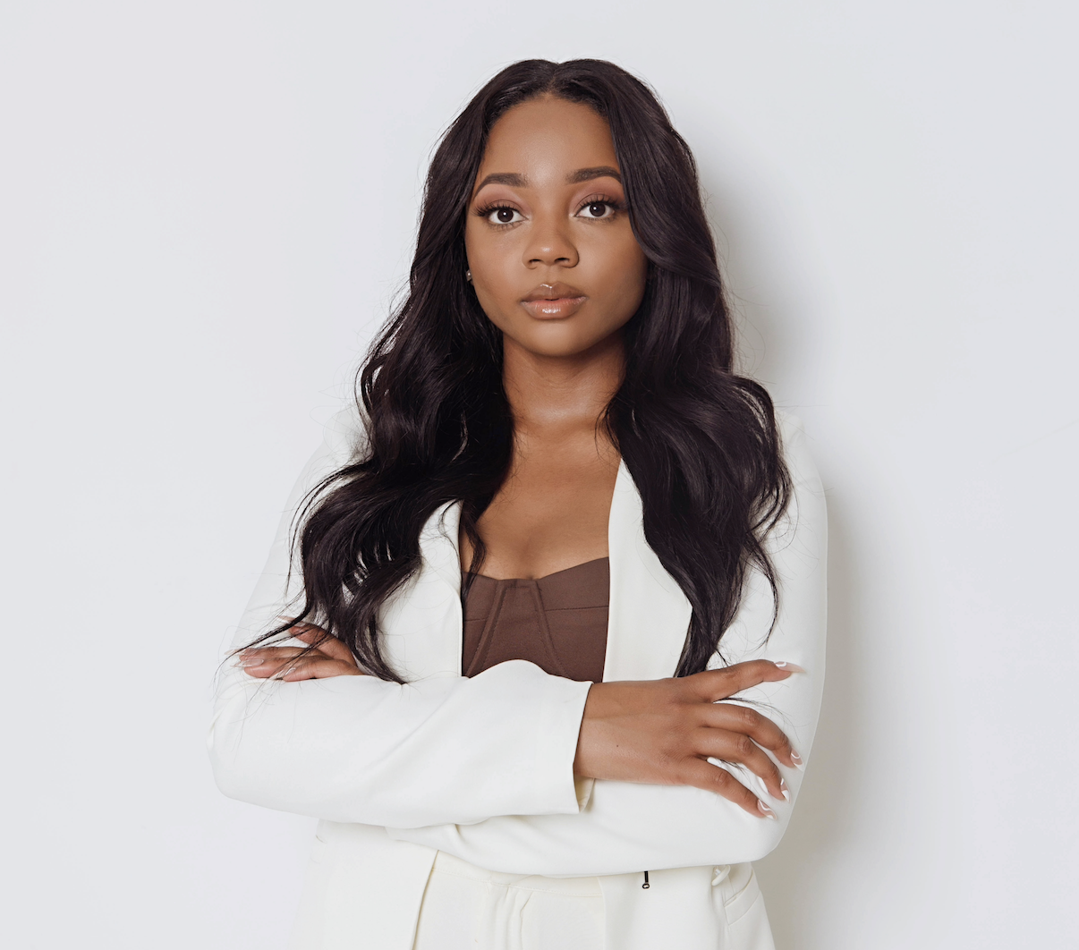 Portrait of a woman with long dark hair wearing a white blazer and a brown top, standing against a plain white background.
