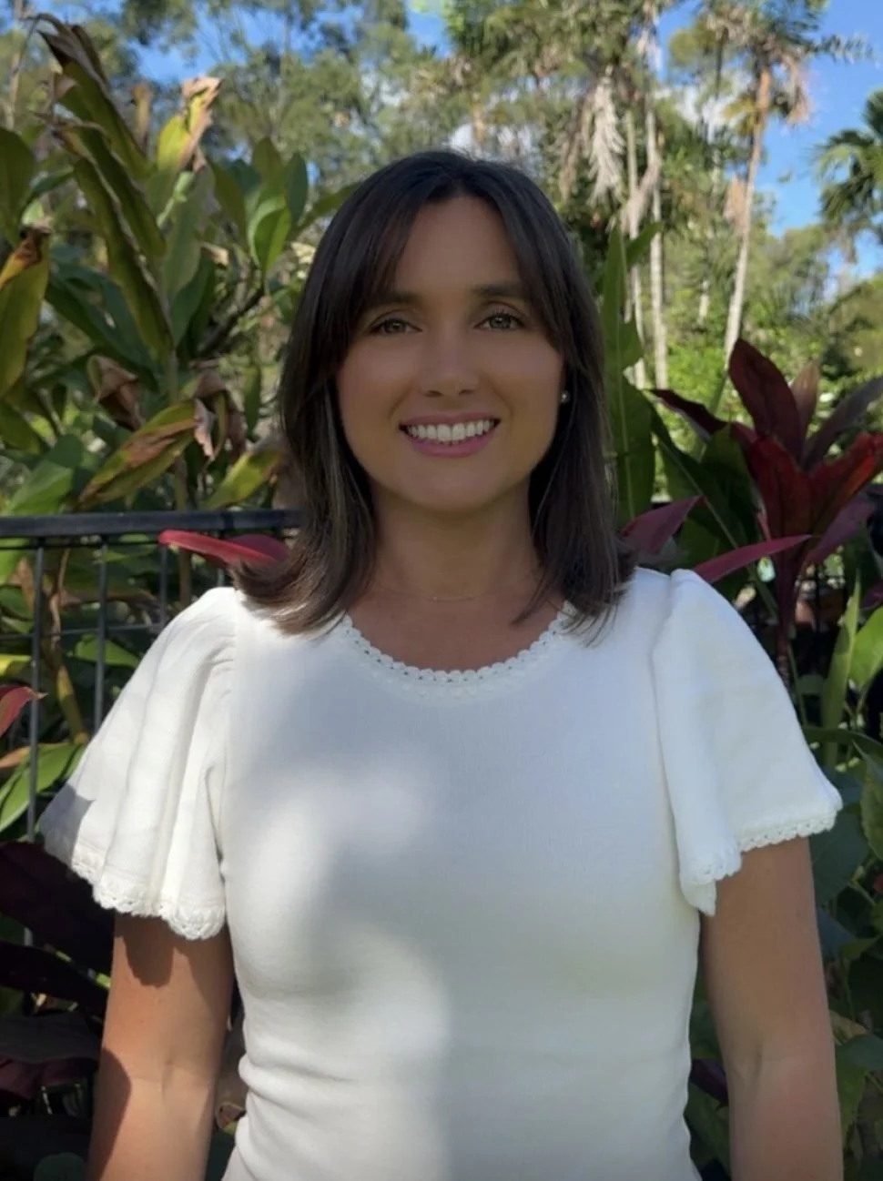 A woman smiling outdoors in front of green, leafy plants and trees.