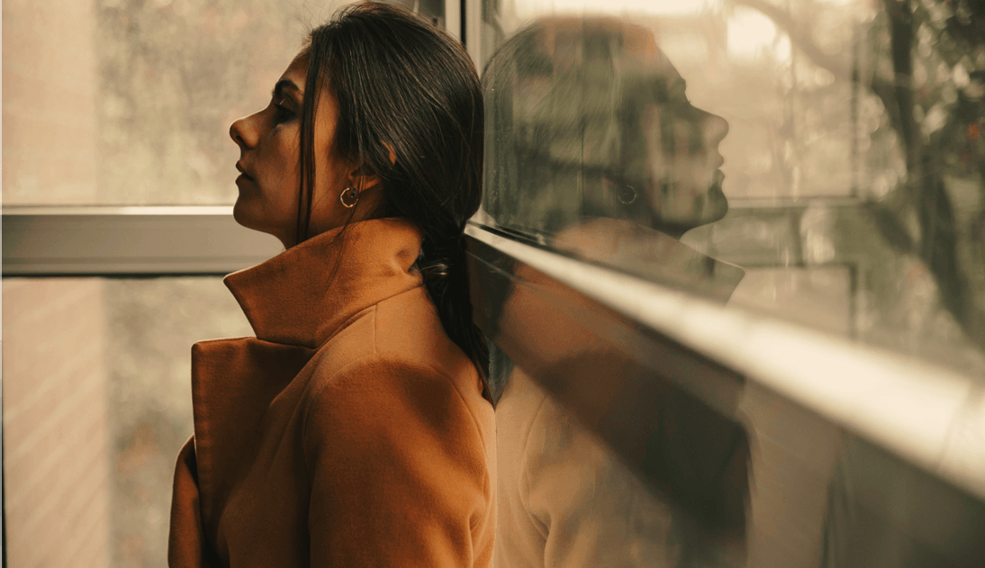 Woman in a brown coat leaning against a glass wall, her reflection visible.
