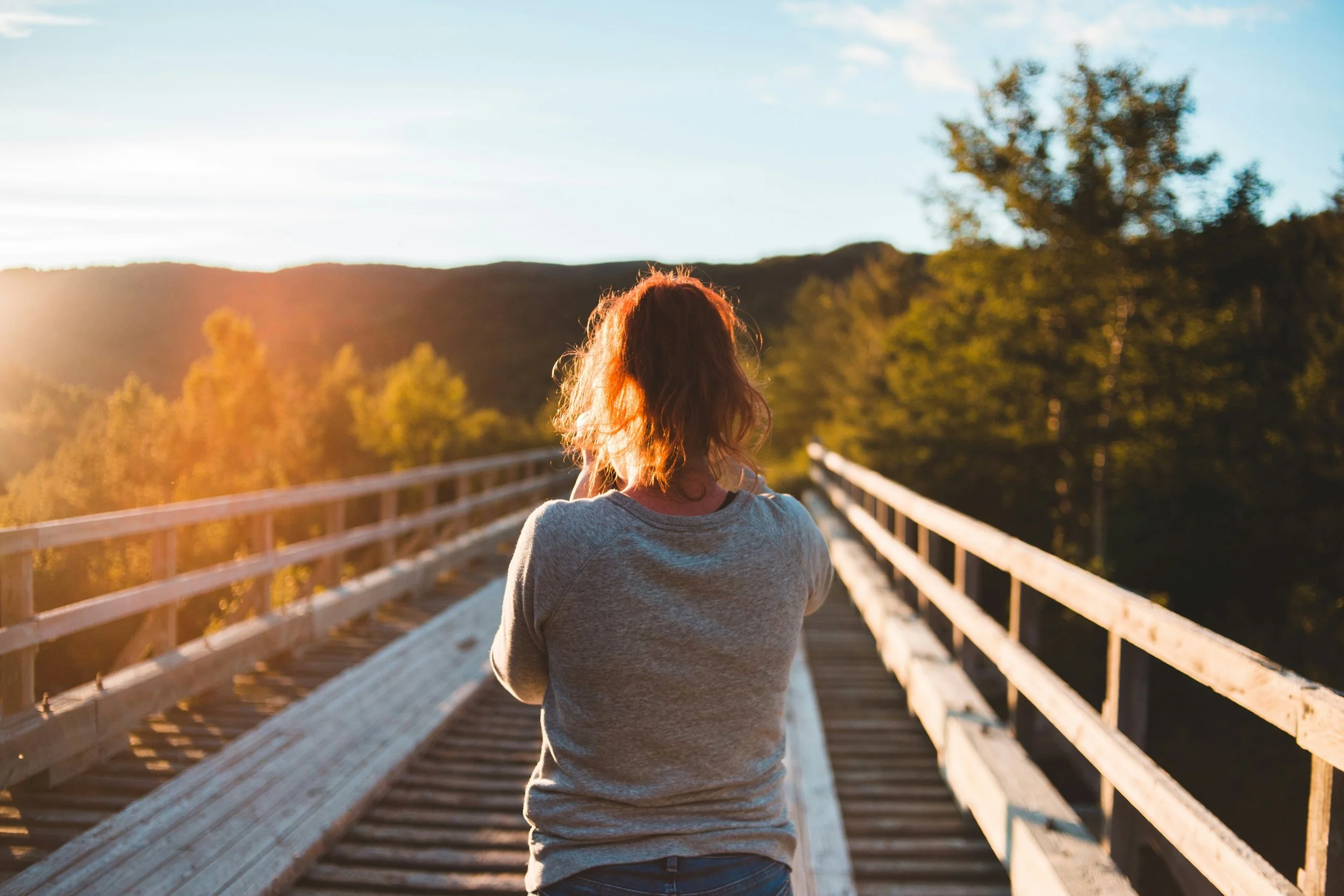 A woman with shoulder-length hair wearing a gray sweatshirt standing on a wooden bridge with railing, looking at a scenic view of trees and hills during sunset.