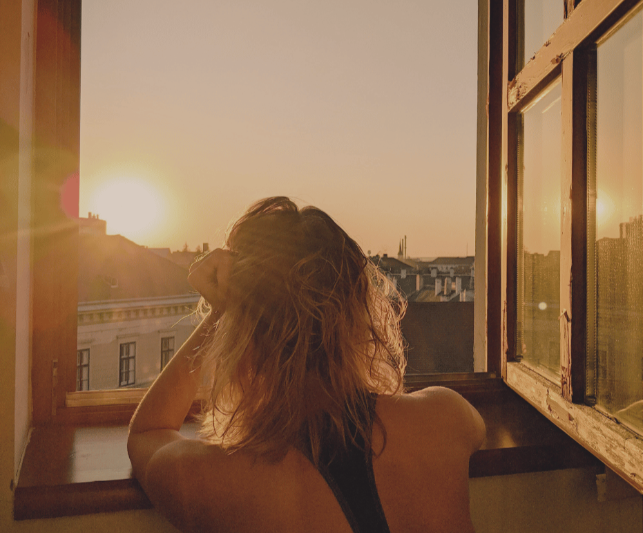 A woman sitting by an open window during sunset, looking outside with her hand resting on her head.