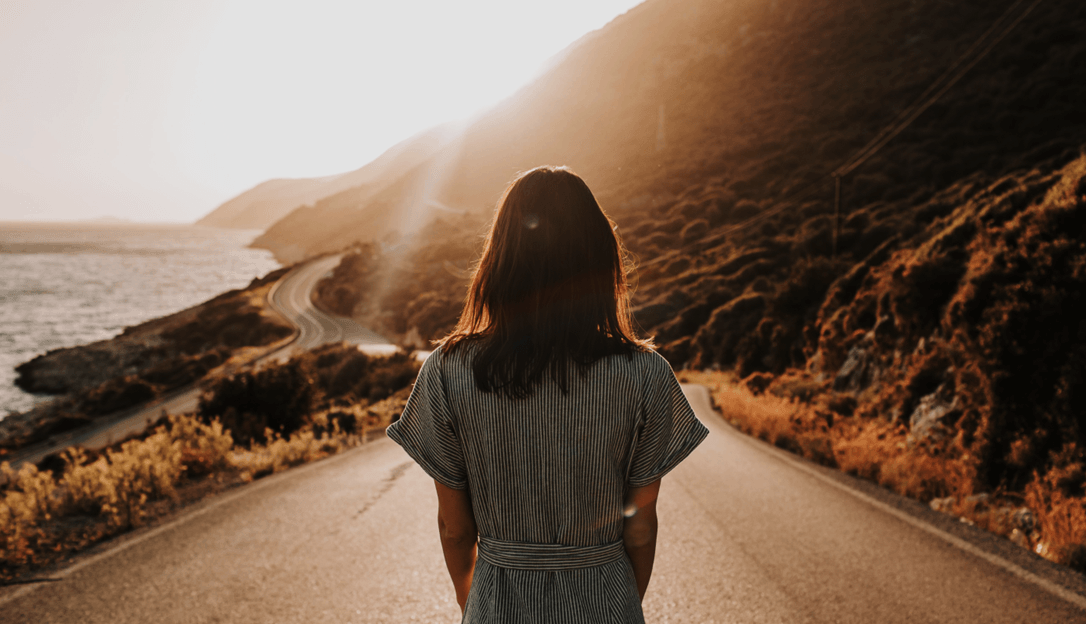 Woman walking down a winding coastal road during sunset with mountains on one side and the ocean on the other.