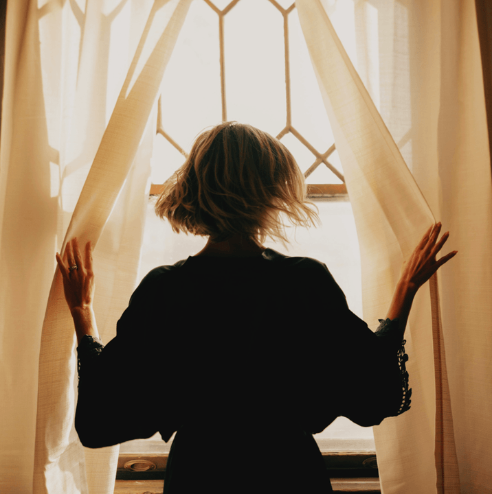 A woman opening curtains to sunlight in front of a window