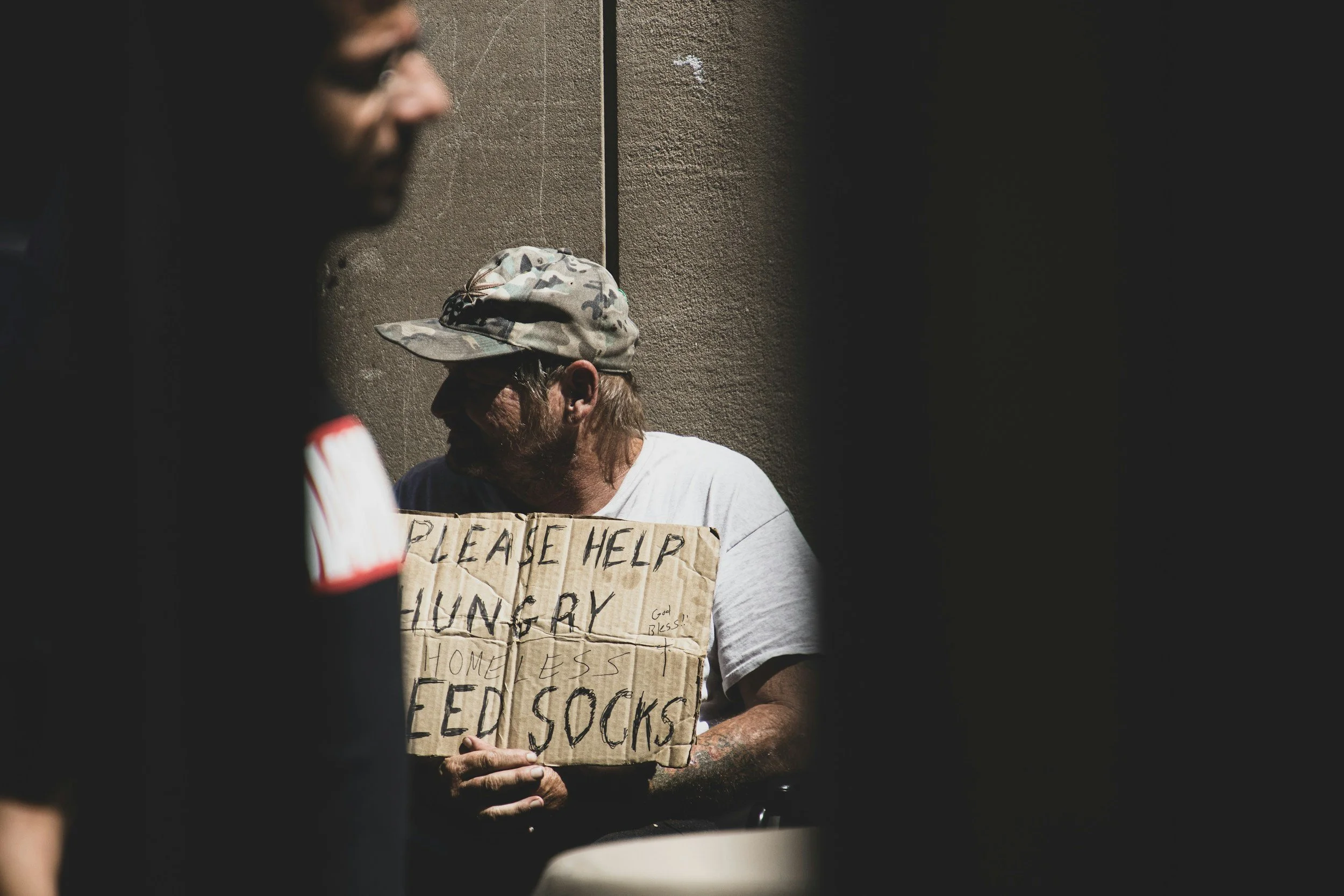 A person holding a cardboard sign that reads "Please help hungry homeless feed Socks", with a camouflage hat, standing against a textured wall. Another person is partially visible in foreground.