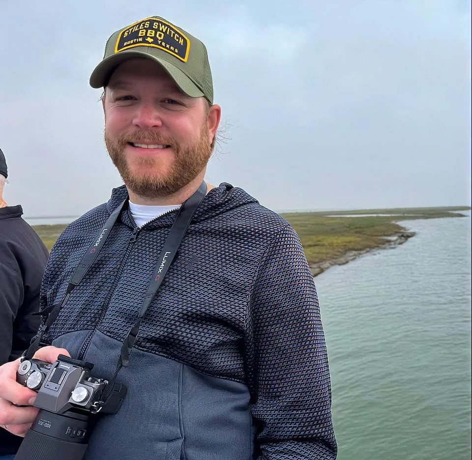 A smiling man holding a camera with a neck strap, standing outdoors near water with marshland in the background.
