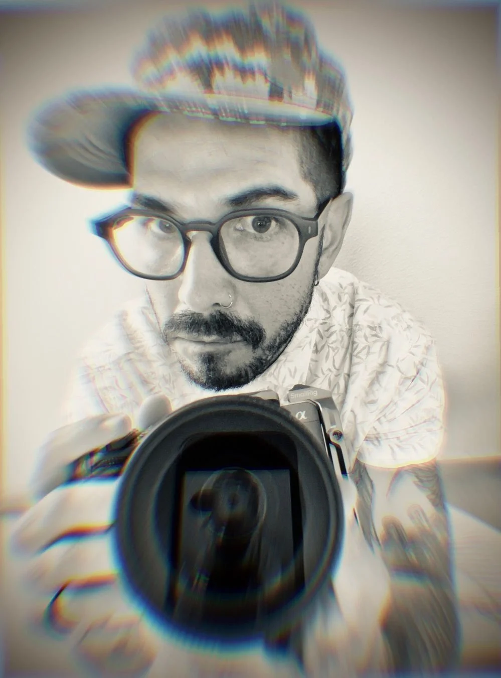 Un hombre con gafas, barba, arete nasal y tatuajes en el brazo, sosteniendo una cámara frente a un espejo, usando una gorra con efecto de distorsión y desenfoque en la imagen.