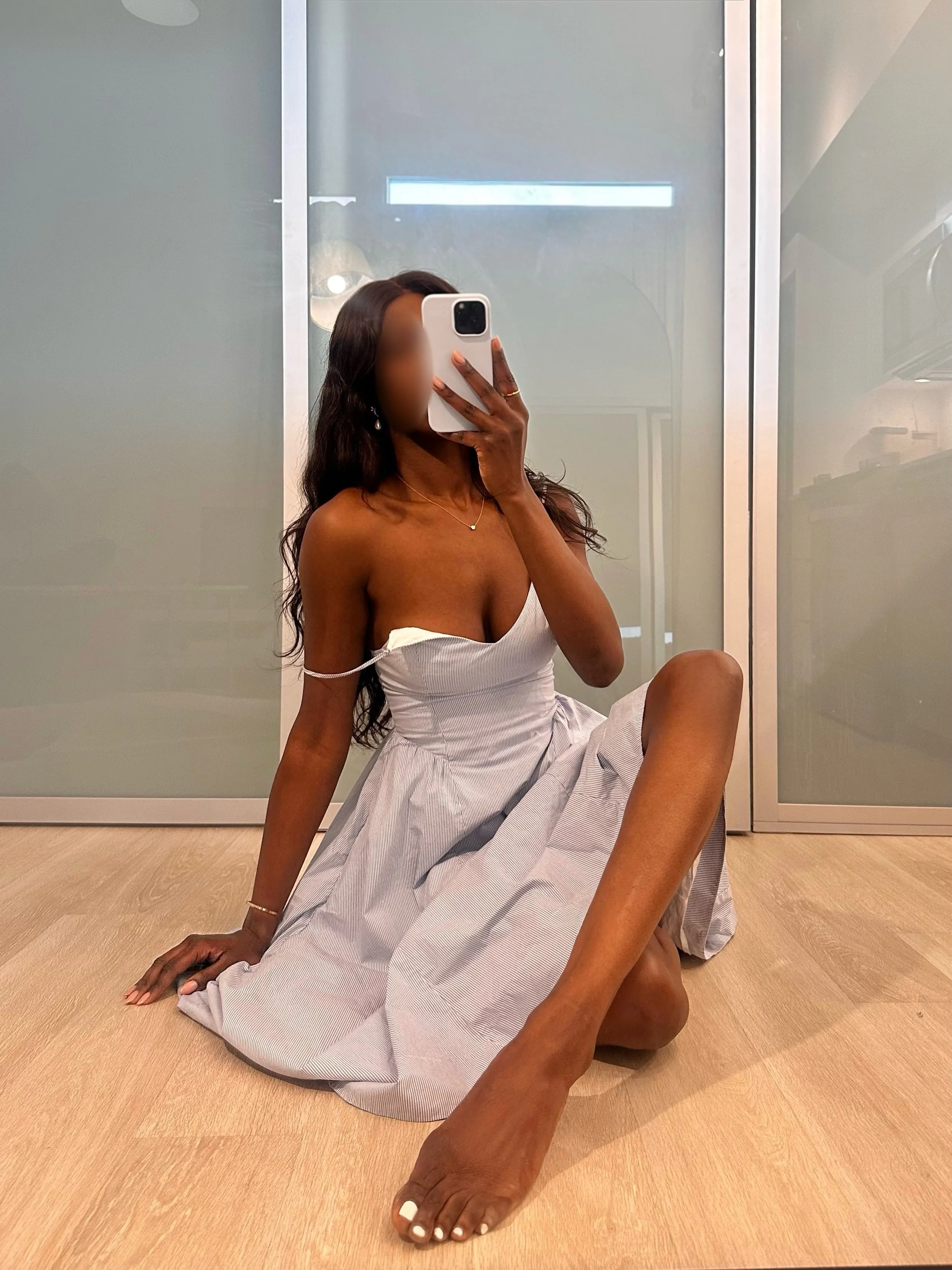 Person sitting on the floor with legs crossed, taking a selfie in front of a mirror, wearing a white dress and jewelry, in a room with wooden flooring and glass doors.