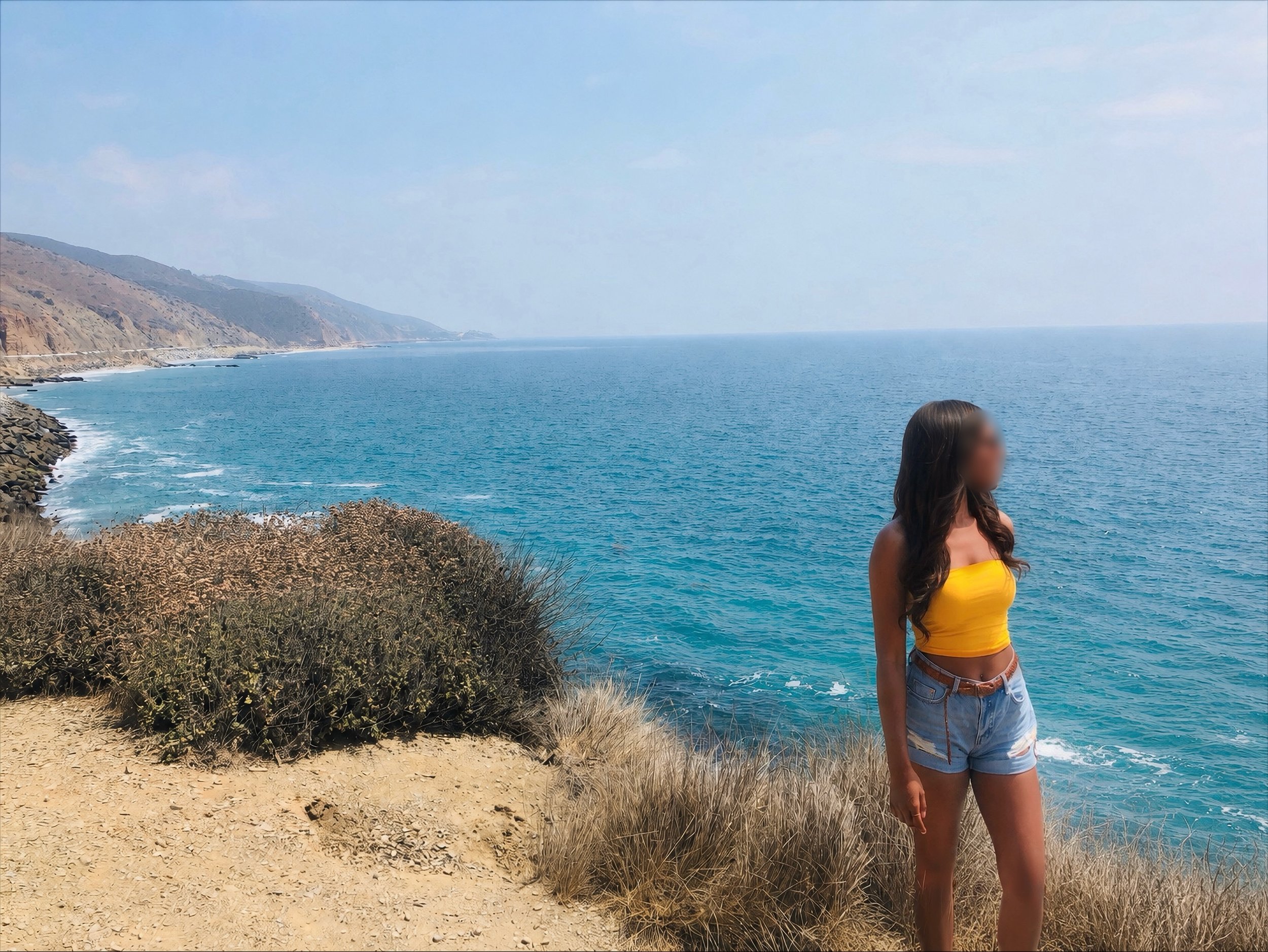 A woman in a yellow crop top and denim shorts standing near the coast, with the ocean, cliffs, and blue sky in the background.