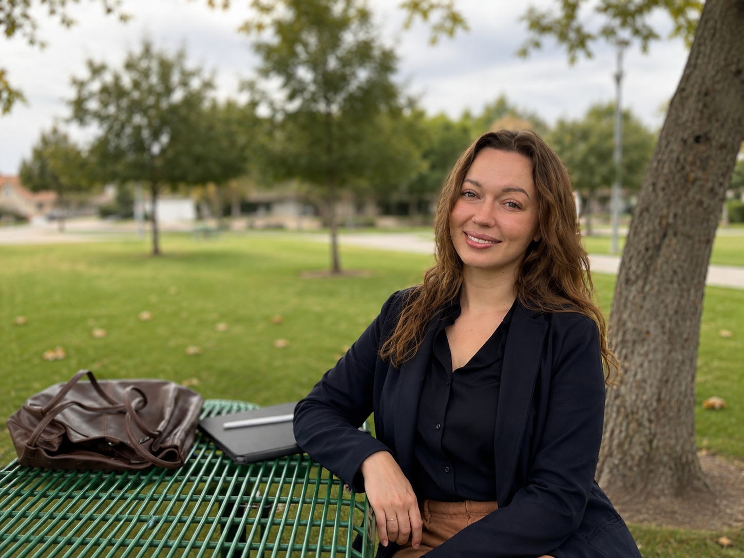 A woman with long curly brown hair and a black blazer sitting at a table in a park, smiling. On the table are a brown purse and a closed tablet. Behind her are trees and a cloudy sky.
