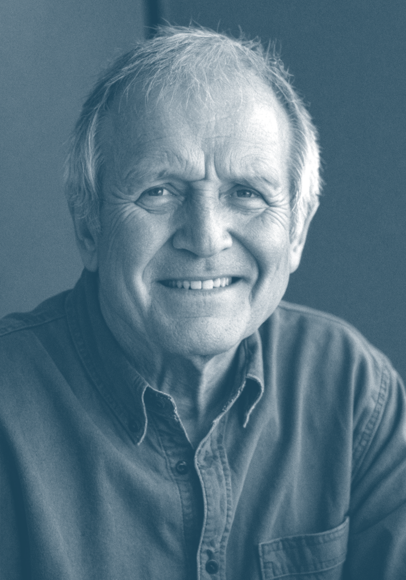 Close-up of an elderly man with white hair and a broad smile, wearing a denim shirt.